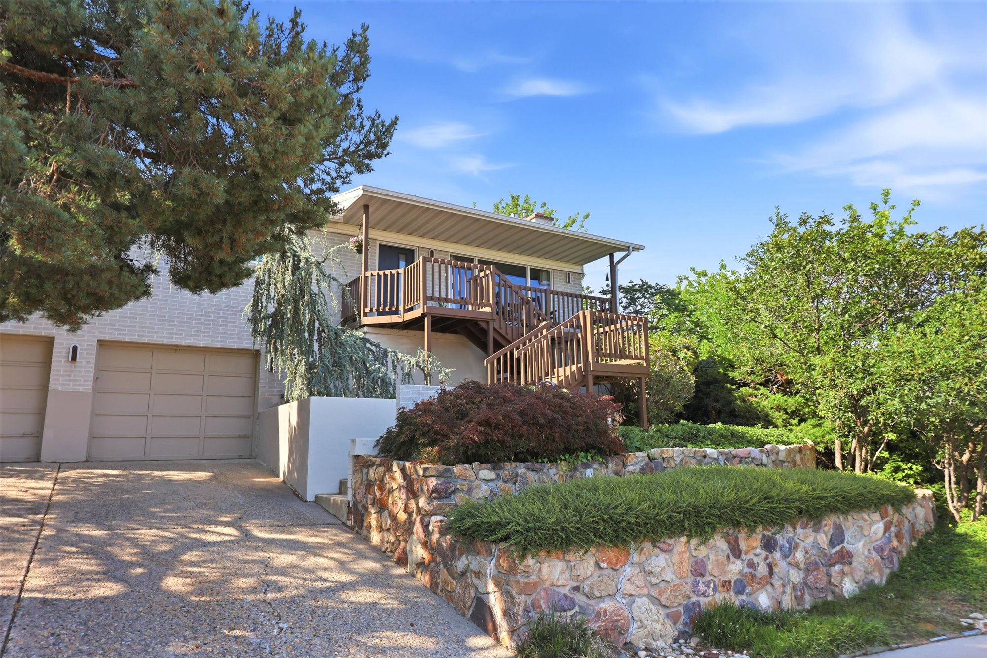 Split-level residence featuring a two-car garage with panel doors, a stone-faced retaining wall, and an elevated wood deck with railing and stairs