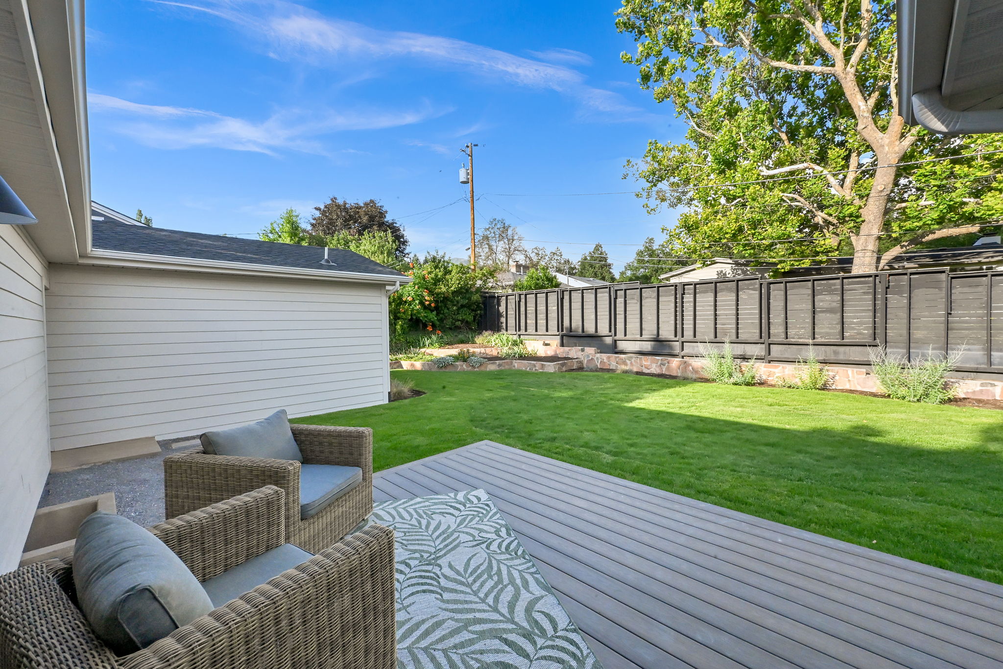 Expansive backyard featuring a gray composite deck, lush green lawn, tiered landscaping with mature plantings, and a horizontal slat privacy fence