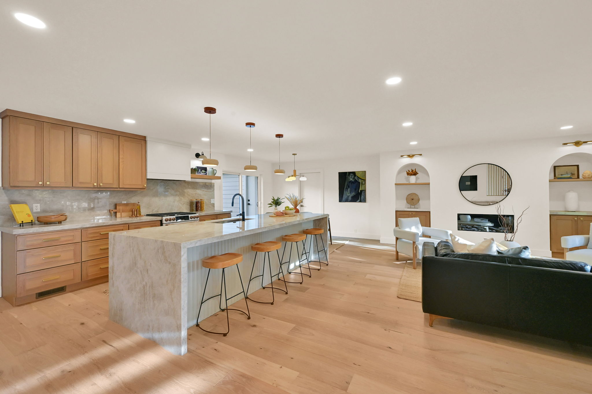 Open-concept living area featuring light wood-finish flooring, a kitchen island with stone waterfall countertop, natural wood cabinetry, a built-in fireplace, and recessed lighting