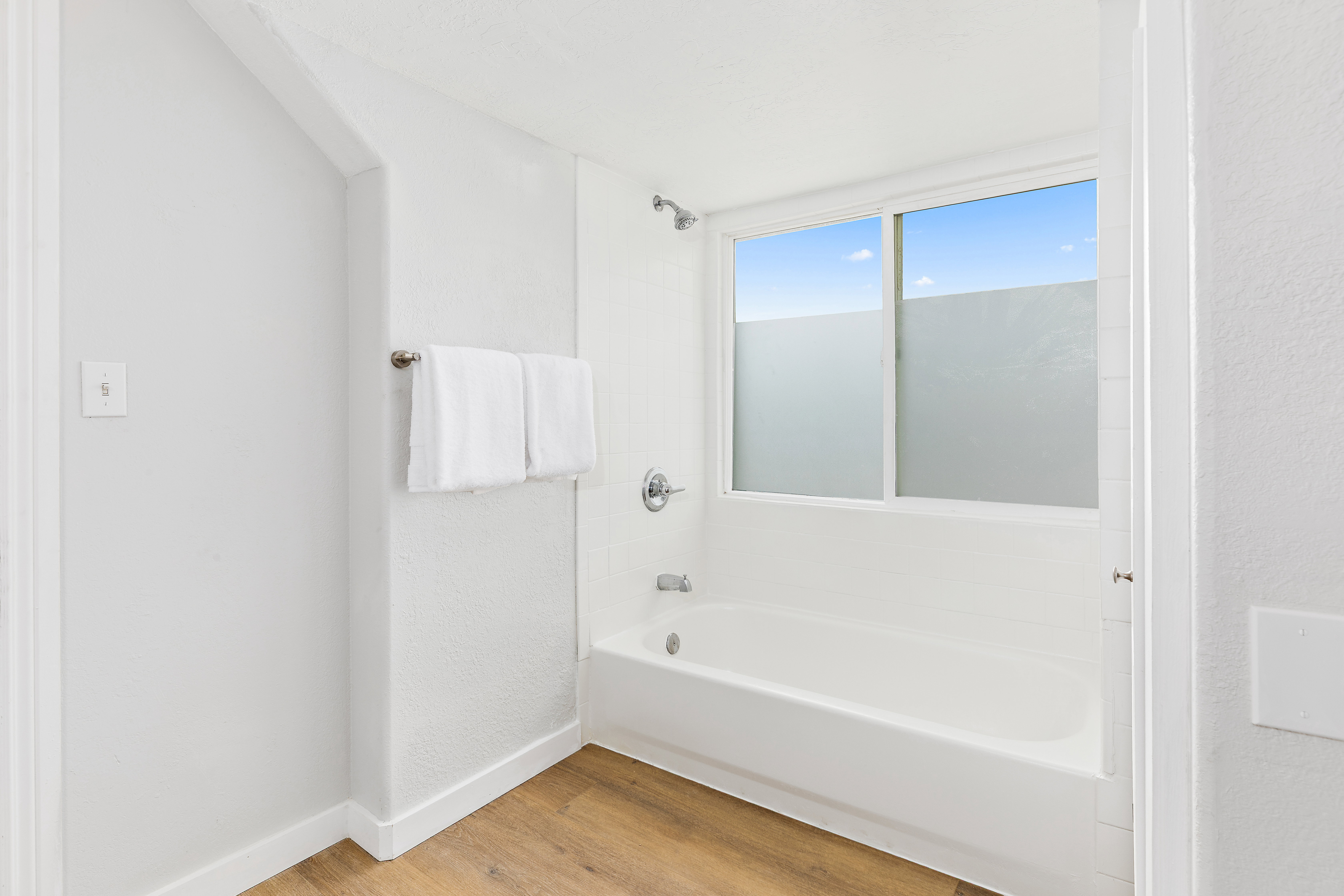 Bathroom featuring a white bathtub, chrome fixtures, a large window with frosted lower panes, white tile surround, and wood-finish flooring