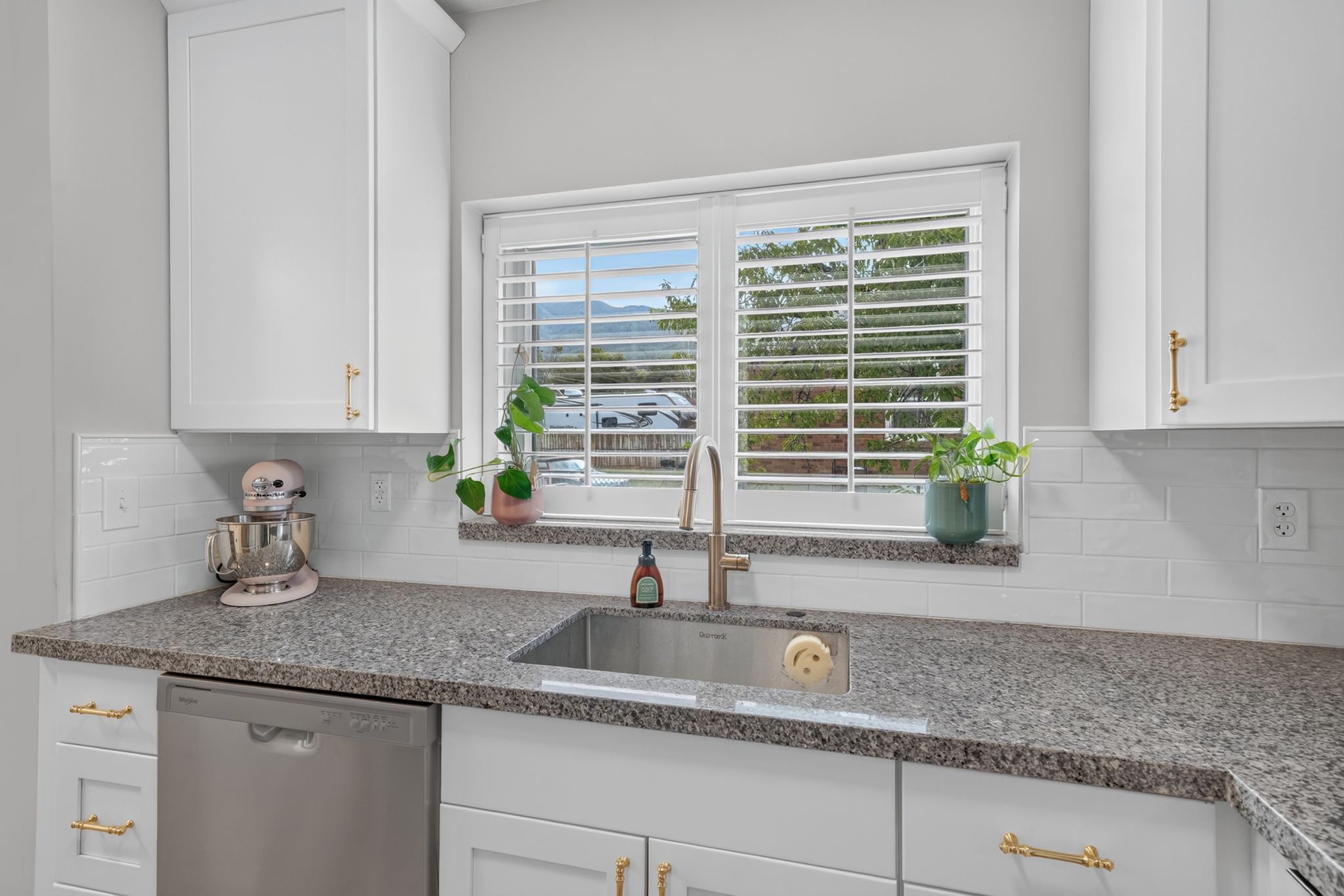 Kitchen sink area featuring a stainless steel undermount sink, granite countertops, white subway tile backsplash, white shaker-style cabinetry with gold hardware, and a stainless steel dishwasher