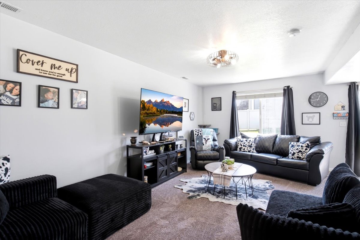 Spacious living area featuring light gray walls, plush carpet flooring, a multi-pane window with dark drapery, a built-in entertainment console, and a contemporary ceiling light fixture