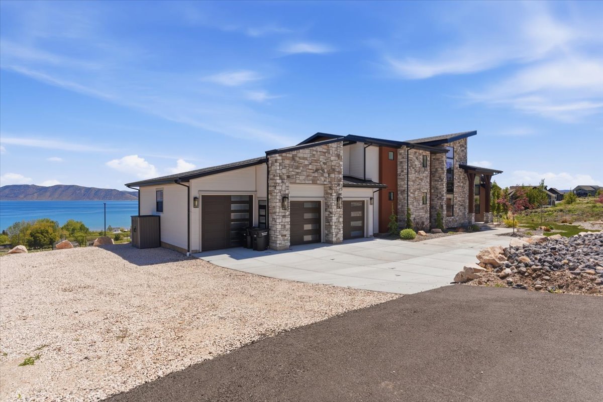 Modern multi-level residence featuring stone and stucco exterior finishes, a multi-car garage with contemporary doors, a concrete driveway, and a gravel parking area