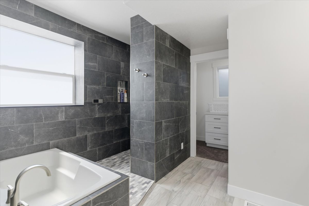 Bathroom featuring a white soaking tub with brushed nickel fixtures, dark gray rectangular wall tiling, a walk-in shower with mosaic tile flooring, and light-toned wood-finish floor tiles