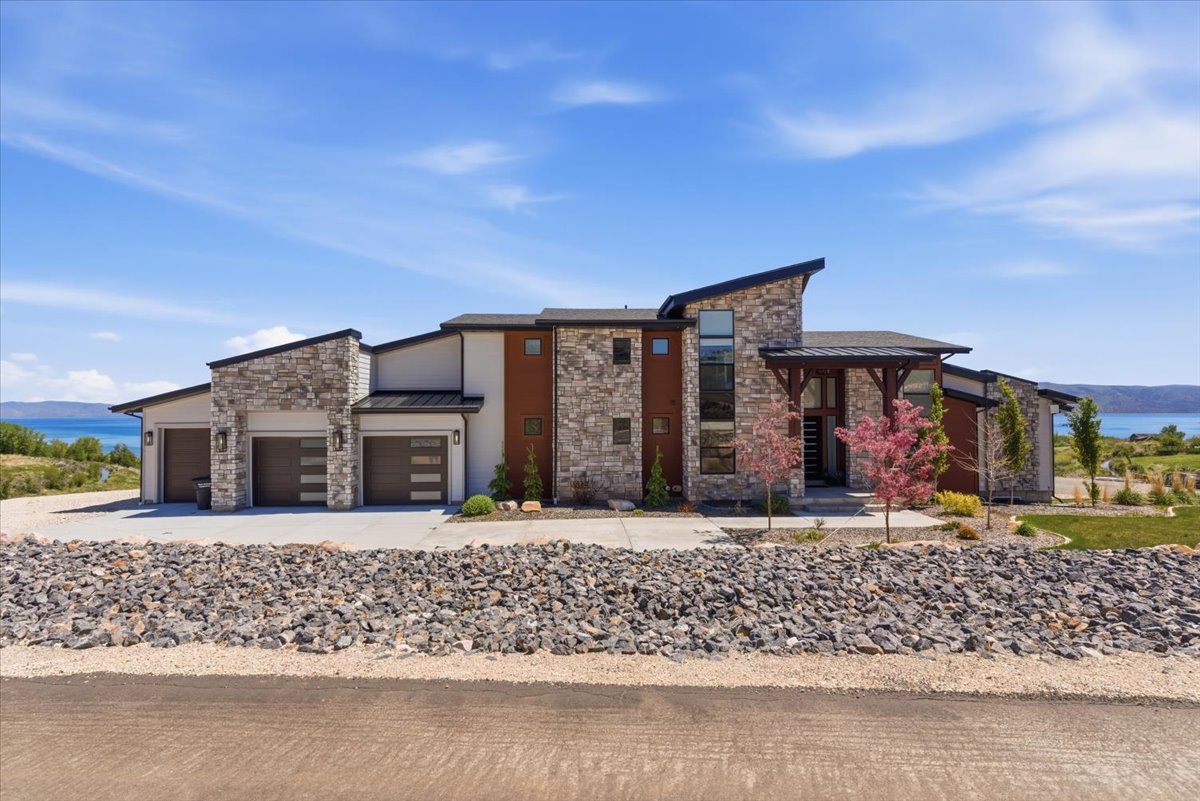 Contemporary architectural design featuring stone and stucco finishes, multiple garage bays with modern doors, a prominent two-story window, a covered entryway with timber accents, and a landscaped front yard with ornamental trees