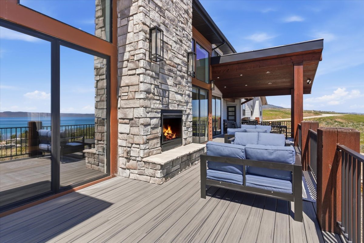Expansive outdoor deck featuring a stone-clad fireplace, covered patio area with recessed lighting, full glass exterior walls, and a composite deck surface with metal railings