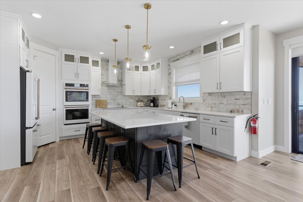 Contemporary kitchen featuring a large central island with seating, white shaker cabinetry, stainless steel appliances, wood-finish flooring, and a subway tile backsplash
