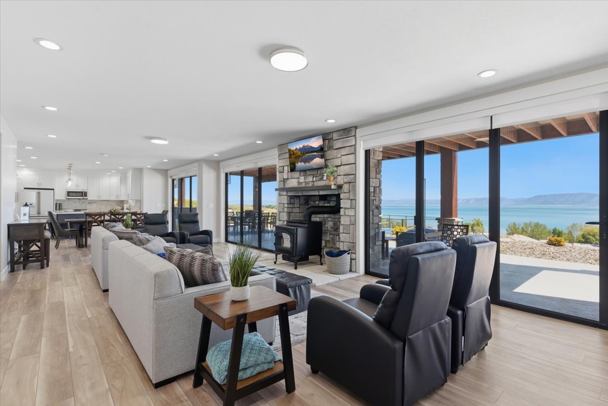 Spacious living area featuring expansive glass sliding doors, a stone-clad fireplace with a wood-burning stove, light wood-finish flooring, recessed lighting, and a contemporary kitchen with white cabinetry and stainless steel appliances