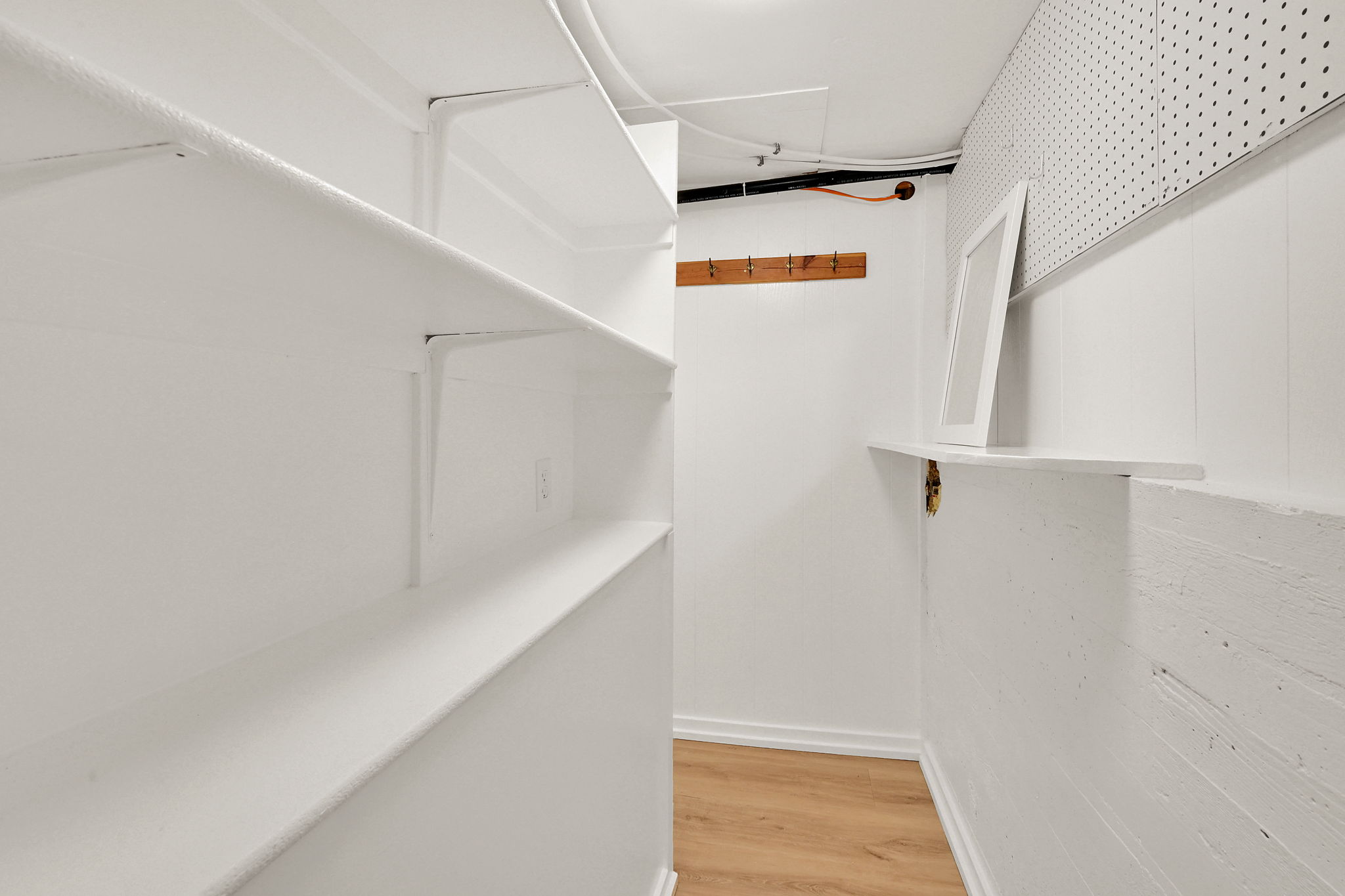 Dedicated storage area featuring extensive built-in shelving, durable wood-finish flooring, a wall-mounted coat rack, a wall-mounted shelf, and pegboard paneling