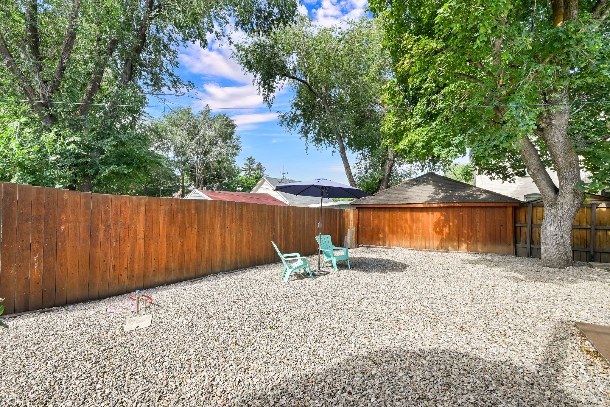 Expansive gravel yard featuring a wood privacy fence, mature trees, and a detached wooden garage with a shingled roof