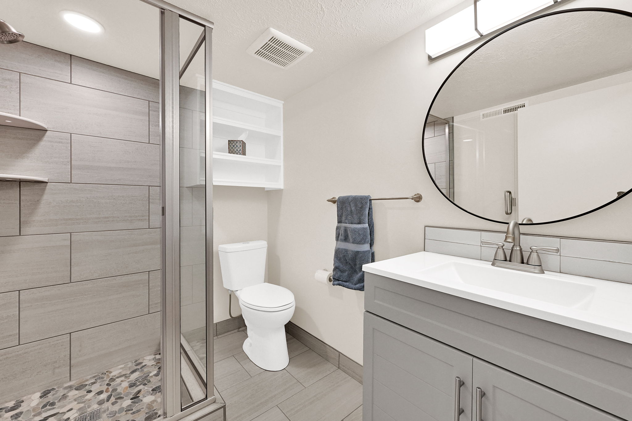Contemporary bathroom featuring a glass-enclosed shower with large format tile surround and pebble floor, a single vanity with a white countertop and grey cabinetry, a circular wall mirror, and a wall-mounted shelving unit