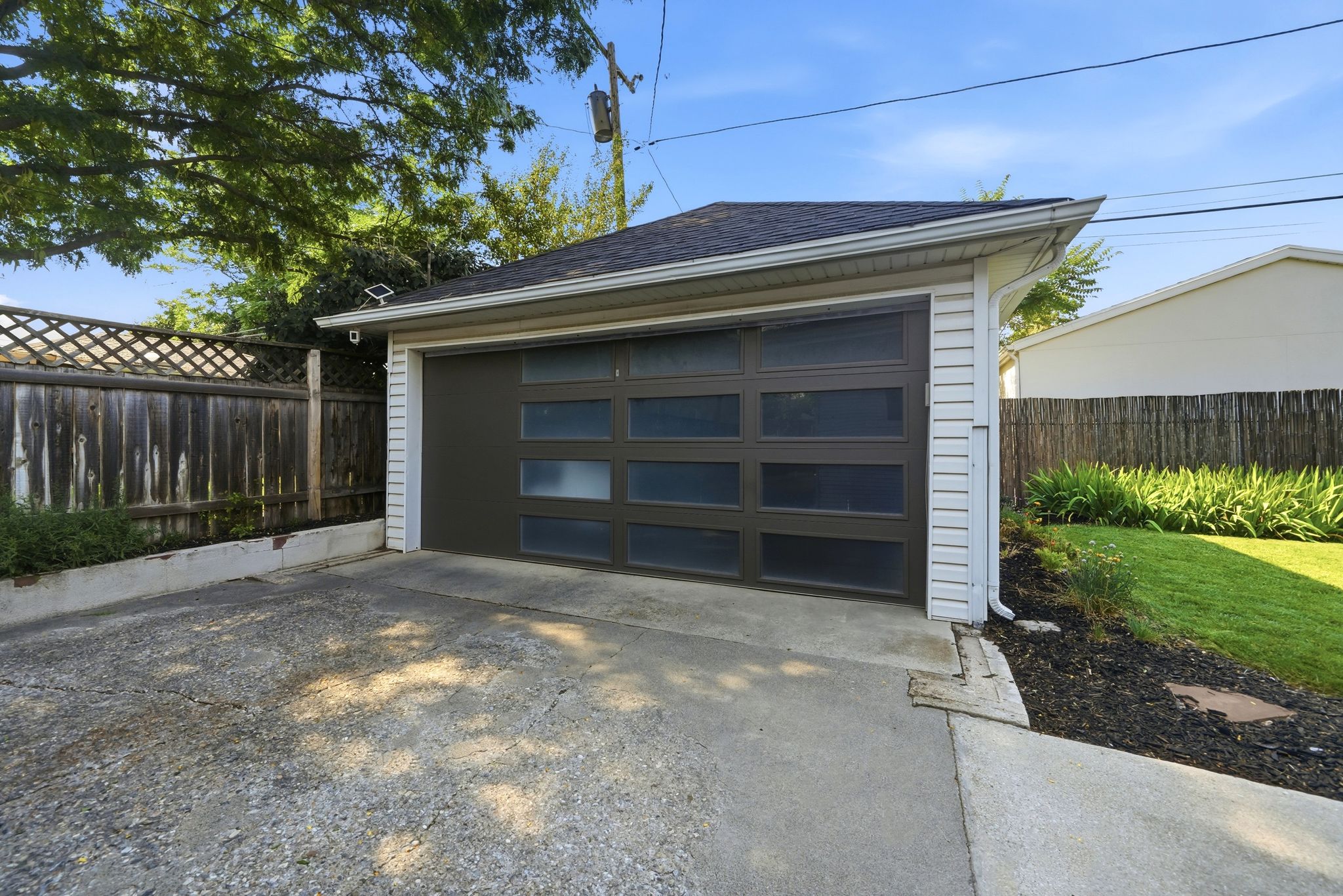 Detached garage with a dark-toned door featuring horizontal frosted glass panels, white siding, and a dark shingled roof