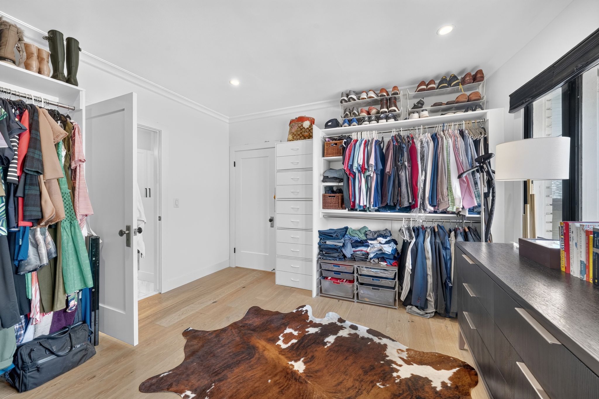 Spacious custom closet featuring light wood-finish flooring, recessed lighting, white built-in shelving, and a large dark wood-finish dresser with metal pulls