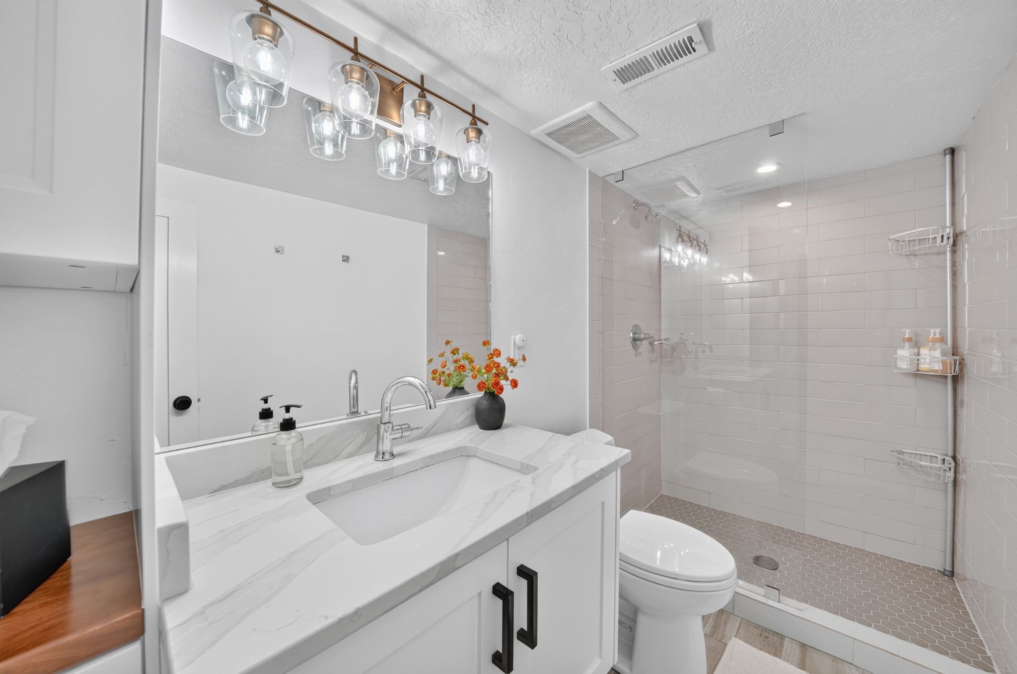 Vanity with a white countertop featuring a single undermount sink, alongside a glass-enclosed shower with tiled walls and a hexagonal mosaic floor