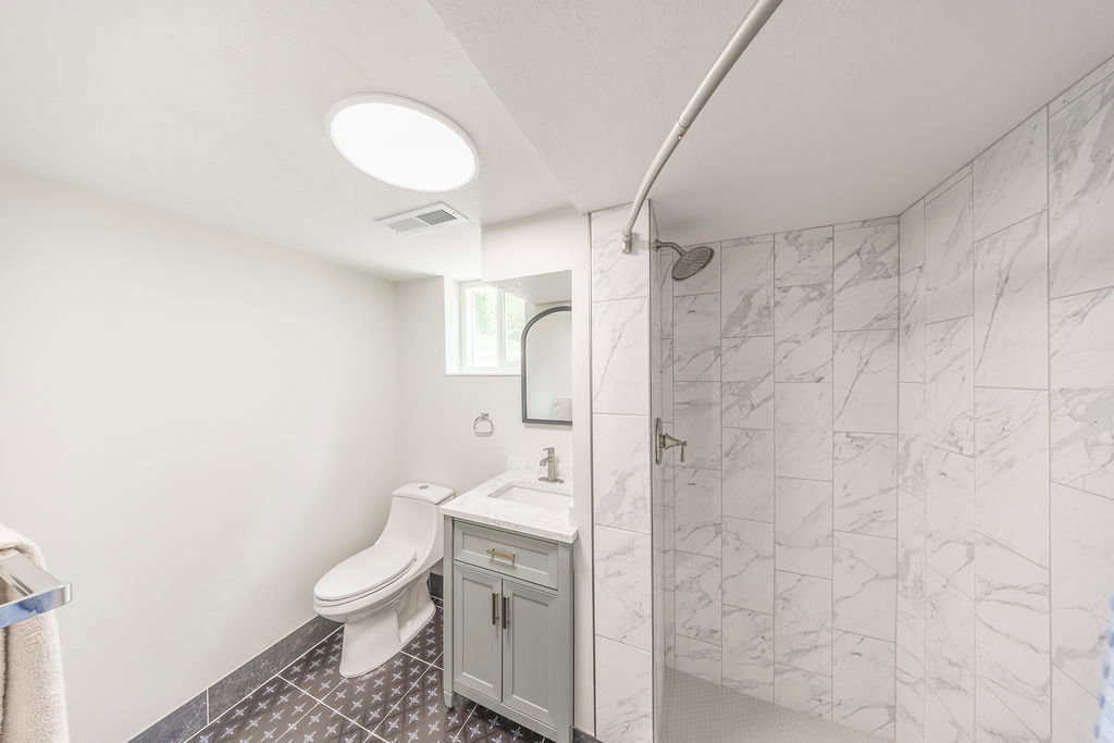 Bathroom with patterned floor tiles, white walls, and a built-in shower featuring large format white marble-look tiles