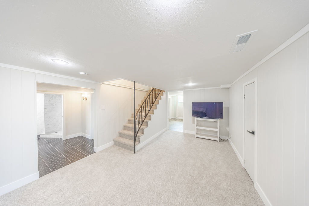 Lower-level living space featuring light-toned carpet, white paneled walls, and a staircase with a black metal railing