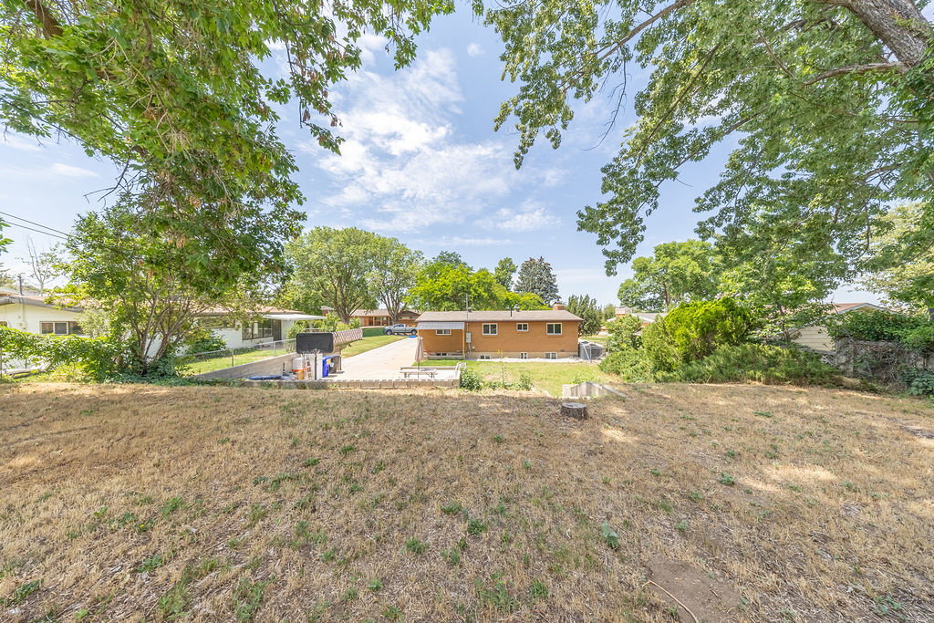 Expansive exterior space featuring a sloped, undeveloped lawn, mature trees, and a distant property with an attached garage