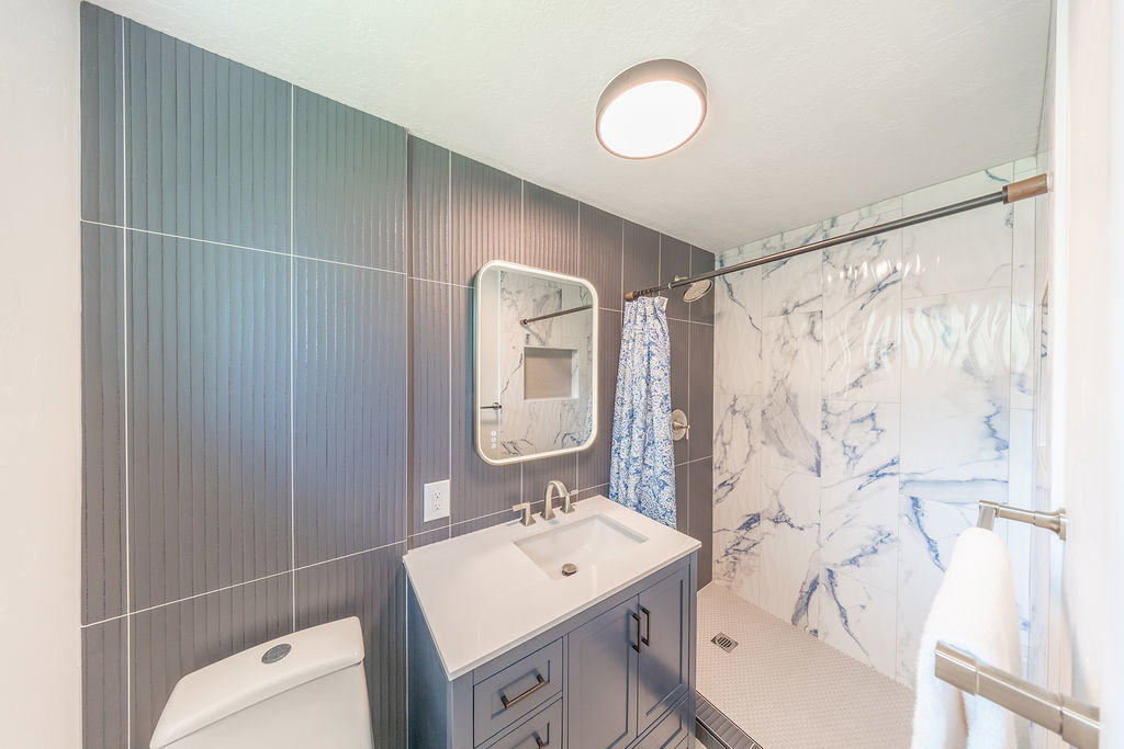 Modern bathroom featuring a single vanity with a white countertop and undermount sink, a rectangular framed mirror, patterned wall tiling, and a walk-in shower with white and blue veined wall panels