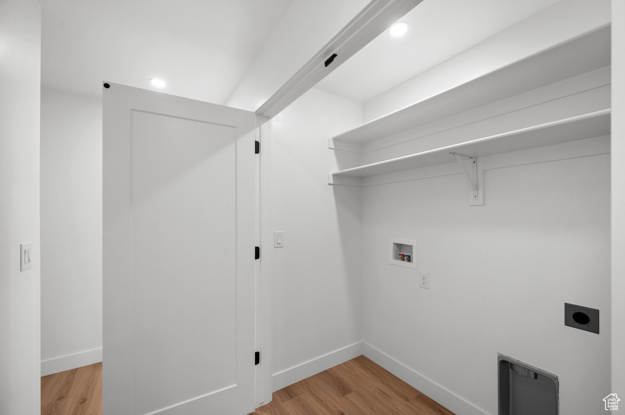 Dedicated utility space featuring two expansive shelves, wood-finish flooring, recessed lighting, and a white flat-panel door with black hardware