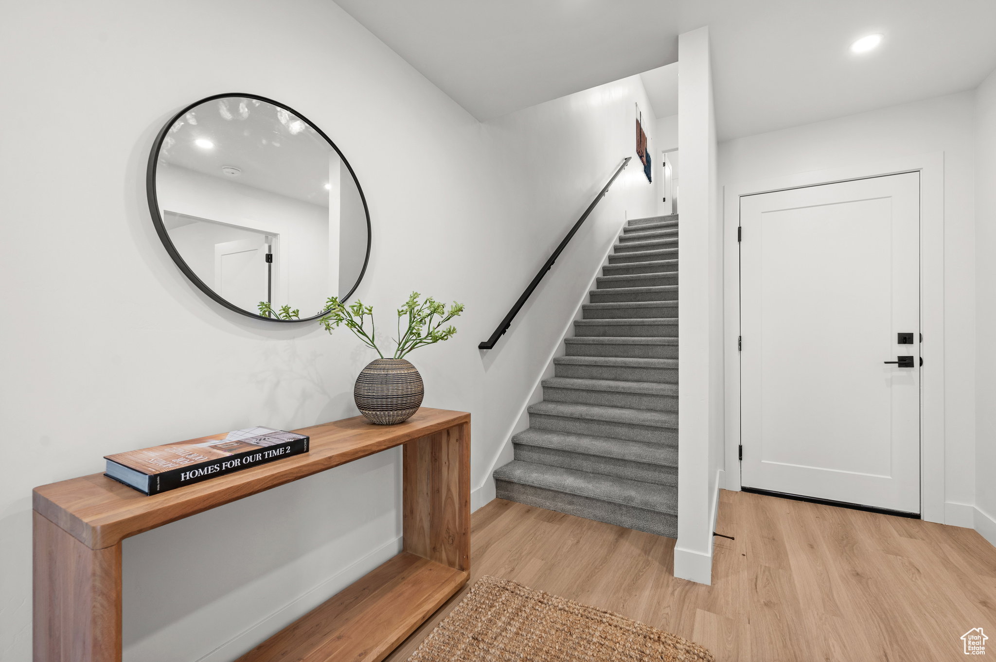 Foyer featuring light wood-finish flooring, white walls, and a gray carpeted staircase with a black handrail