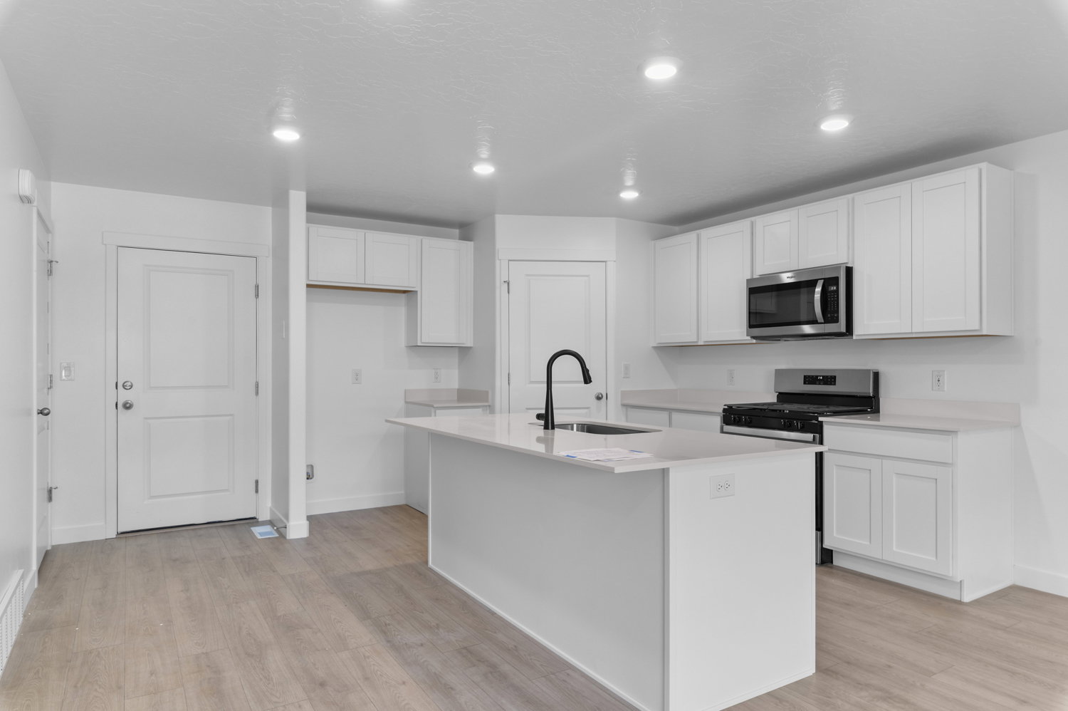 Modern kitchen featuring a central island with an integrated sink, light-toned countertops, white shaker cabinetry, stainless steel appliances, and wood-finish flooring