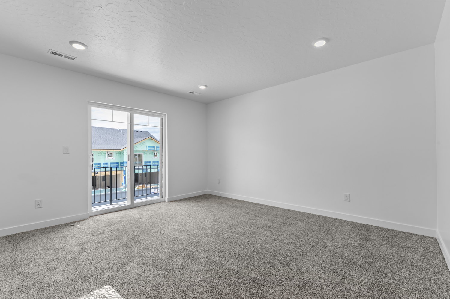 Spacious room featuring new gray carpet, white walls, recessed lighting, and a sliding glass door leading to an exterior balcony with a black metal railing