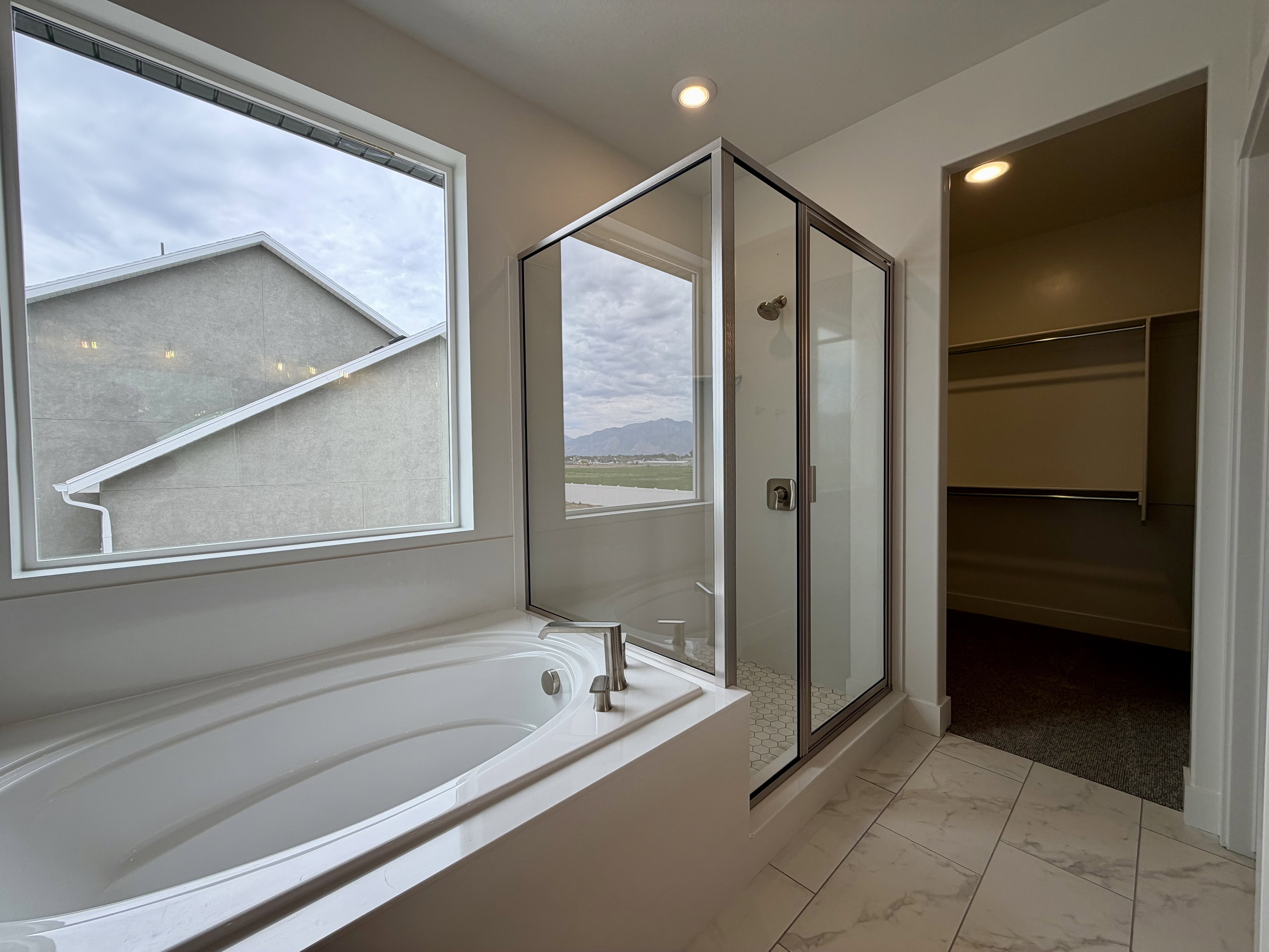 Soaking tub with brushed nickel fixtures, glass-enclosed shower with tiled floor, large window, overhead lighting, and an adjacent walk-in closet with shelving and hanging rods