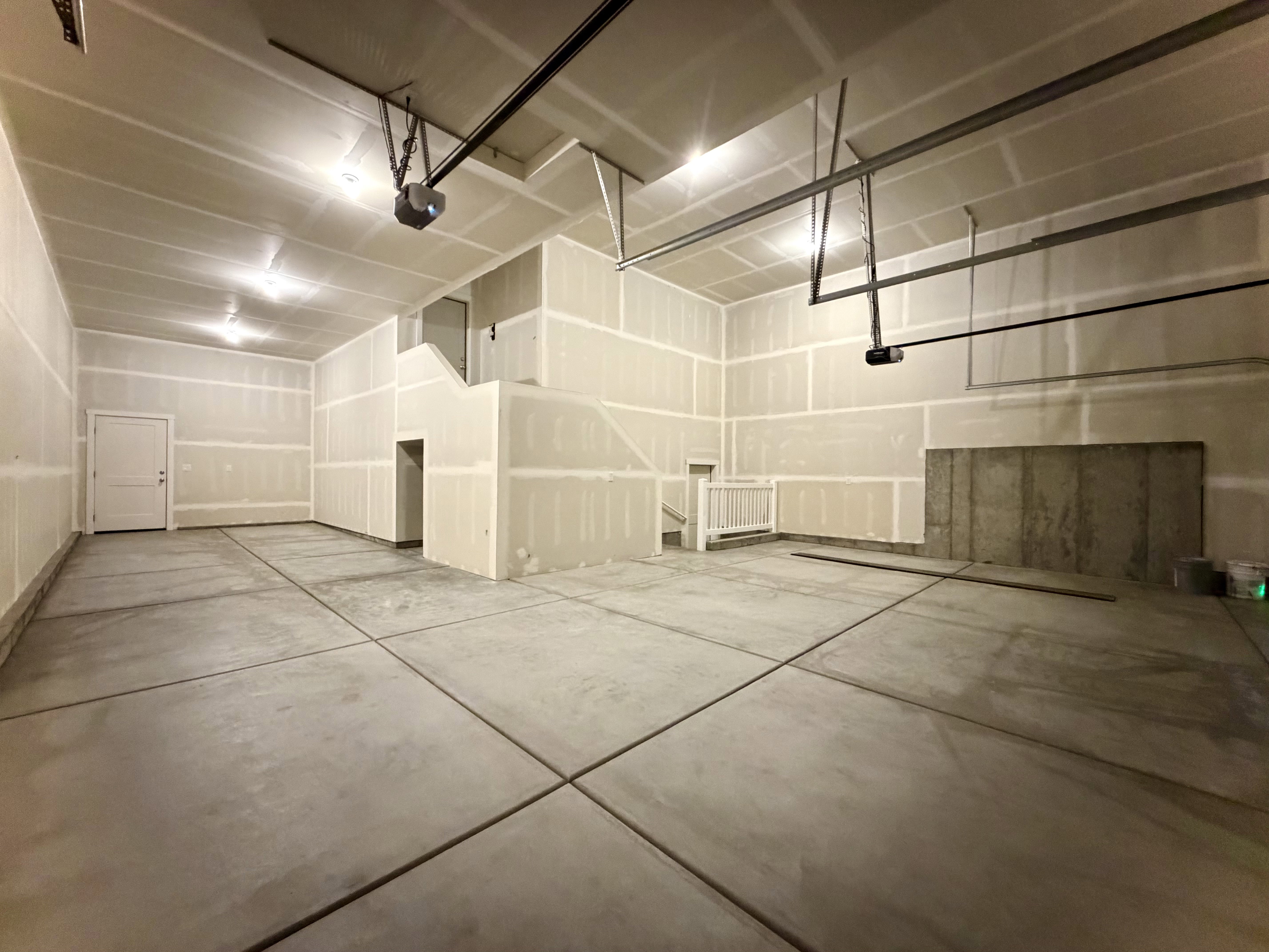 Spacious unfinished garage featuring concrete flooring, unpainted drywall, high ceilings with exposed garage door mechanisms, and a white interior door