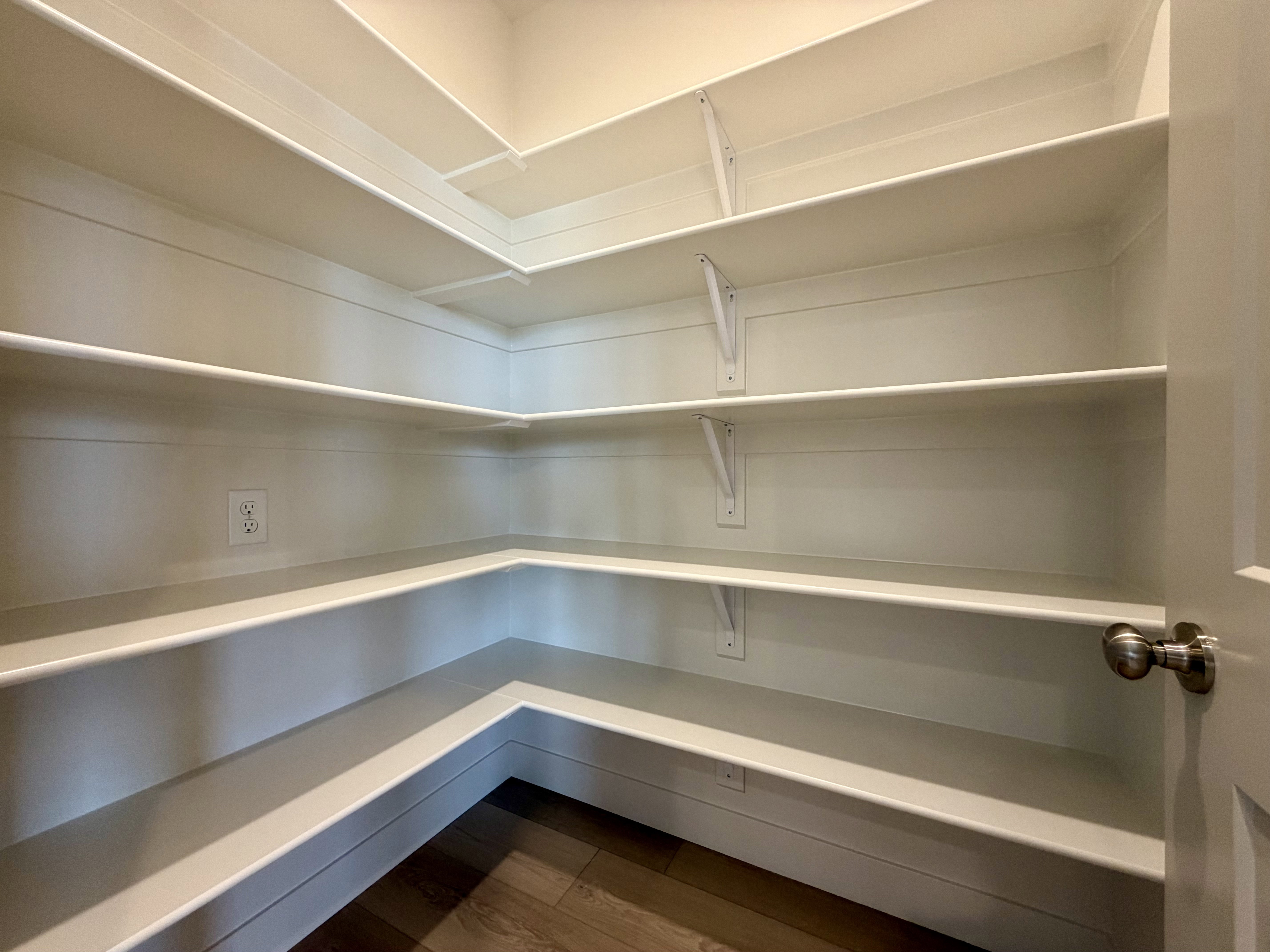 Spacious pantry featuring extensive wrap-around shelving, clean white finishes, wood-finish flooring, and a convenient wall outlet