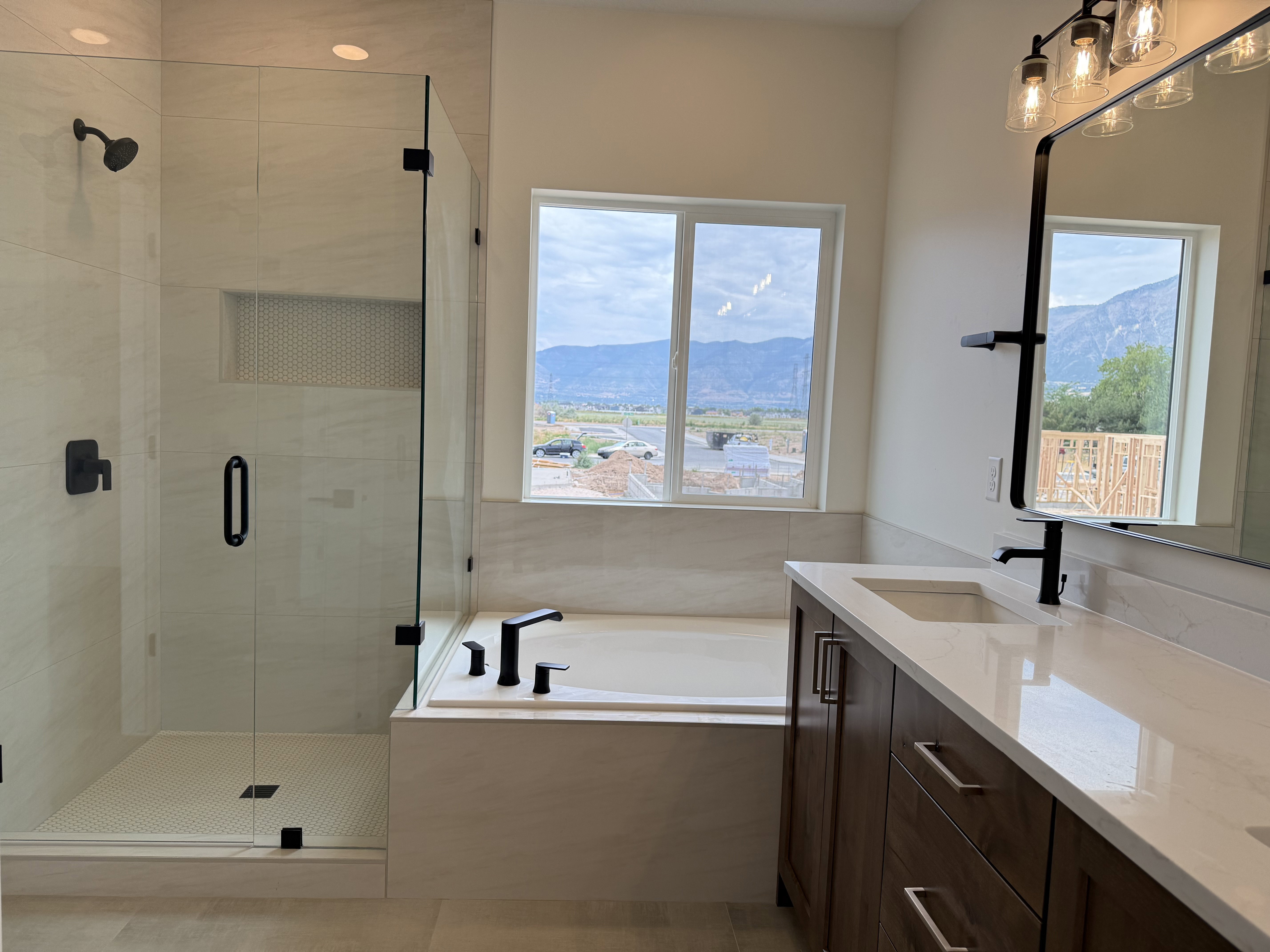 Spacious bathroom featuring a large soaking tub, separate glass-enclosed shower with matte black fixtures, wood-finish vanity with a light-toned countertop, and a large window providing natural light and mountain scenery