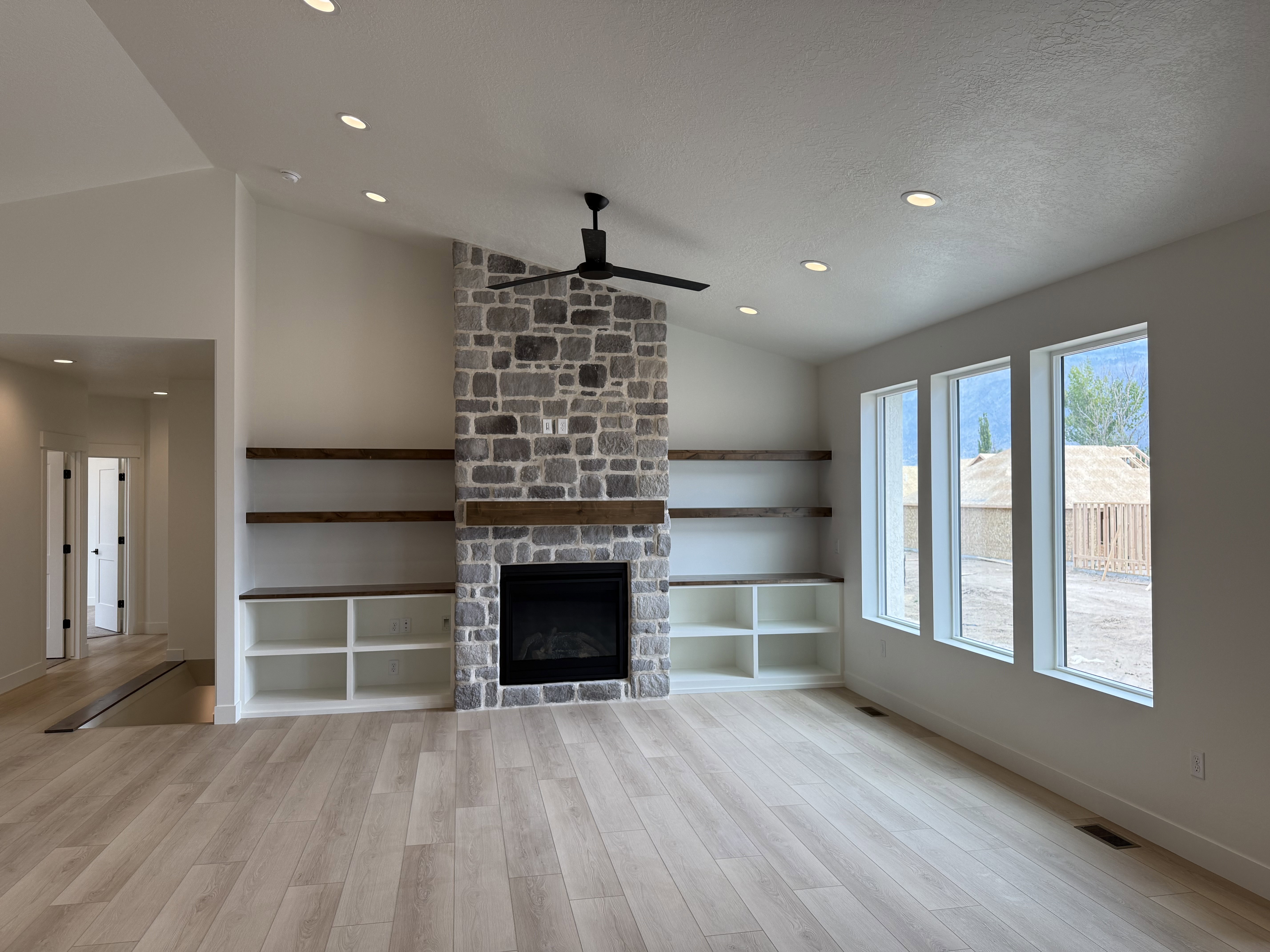 Spacious living area featuring a stone-clad fireplace with a wood mantel, built-in shelving, wood-finish flooring, recessed lighting, and large windows