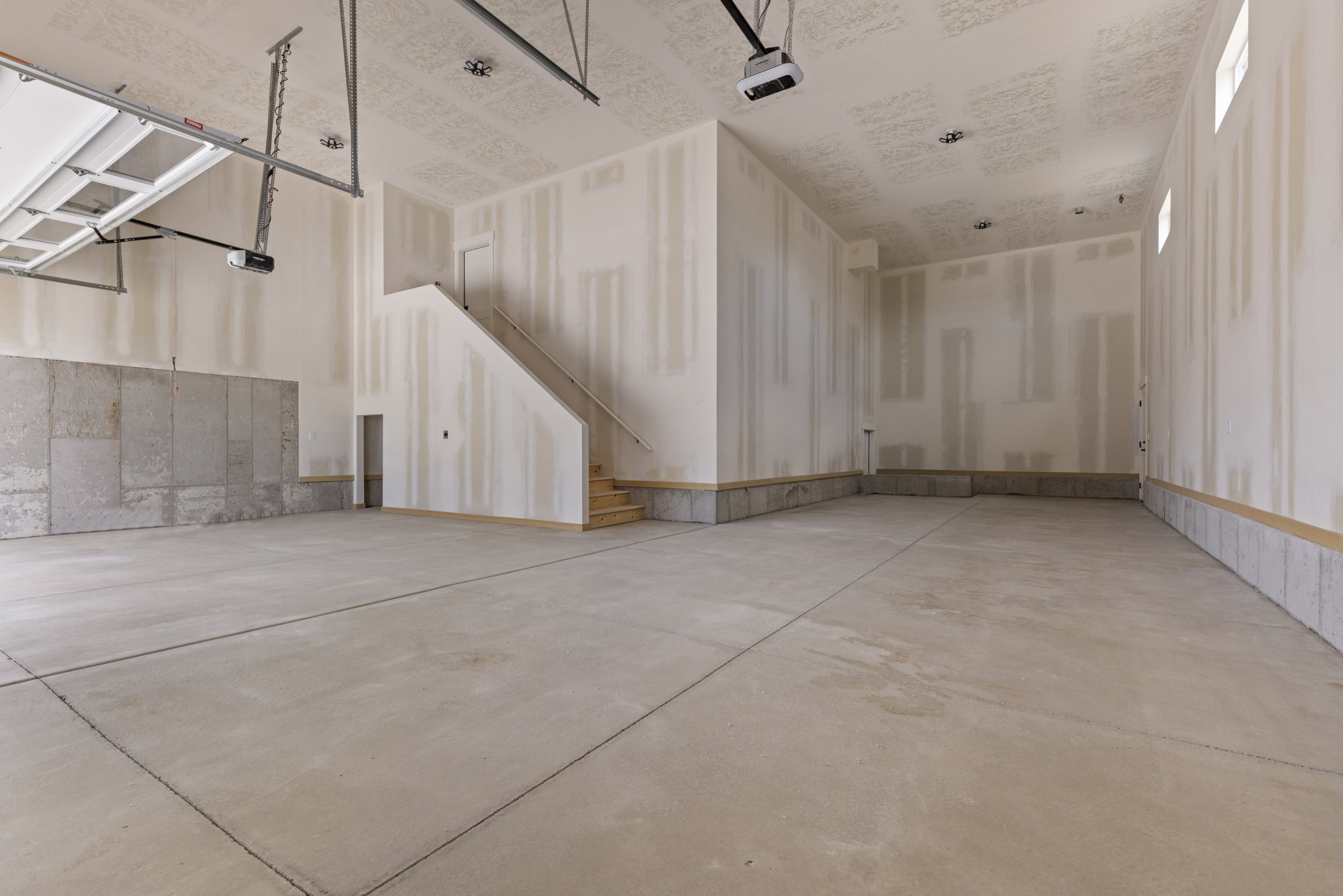Spacious garage with exposed concrete flooring, a partial concrete wall, and an unfinished staircase leading to an upper level