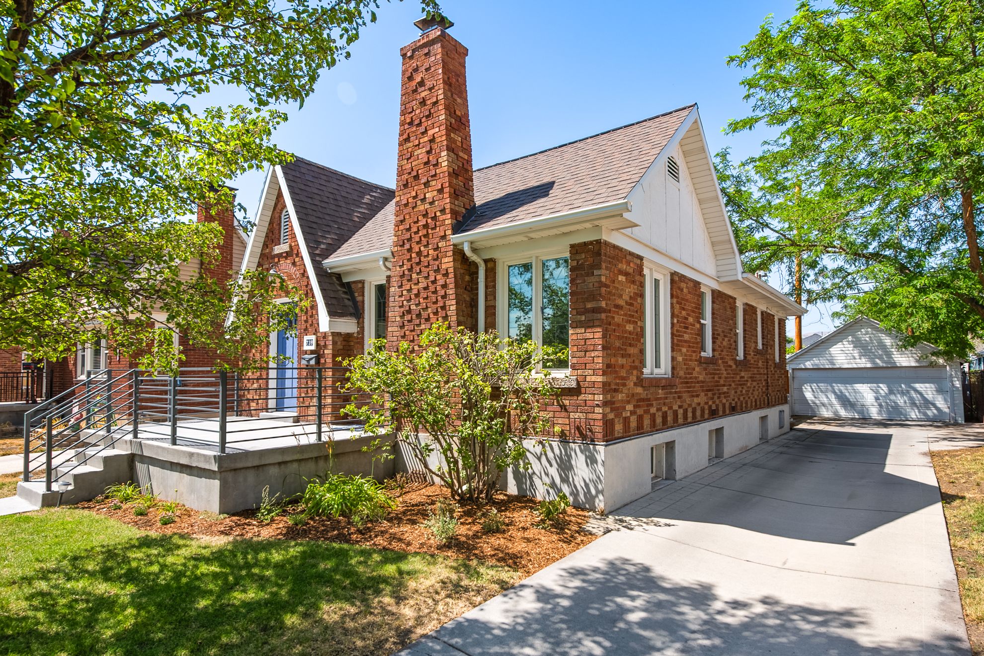 Brick exterior with a prominent chimney, concrete front porch with modern railings, and a side-access driveway leading to a detached garage