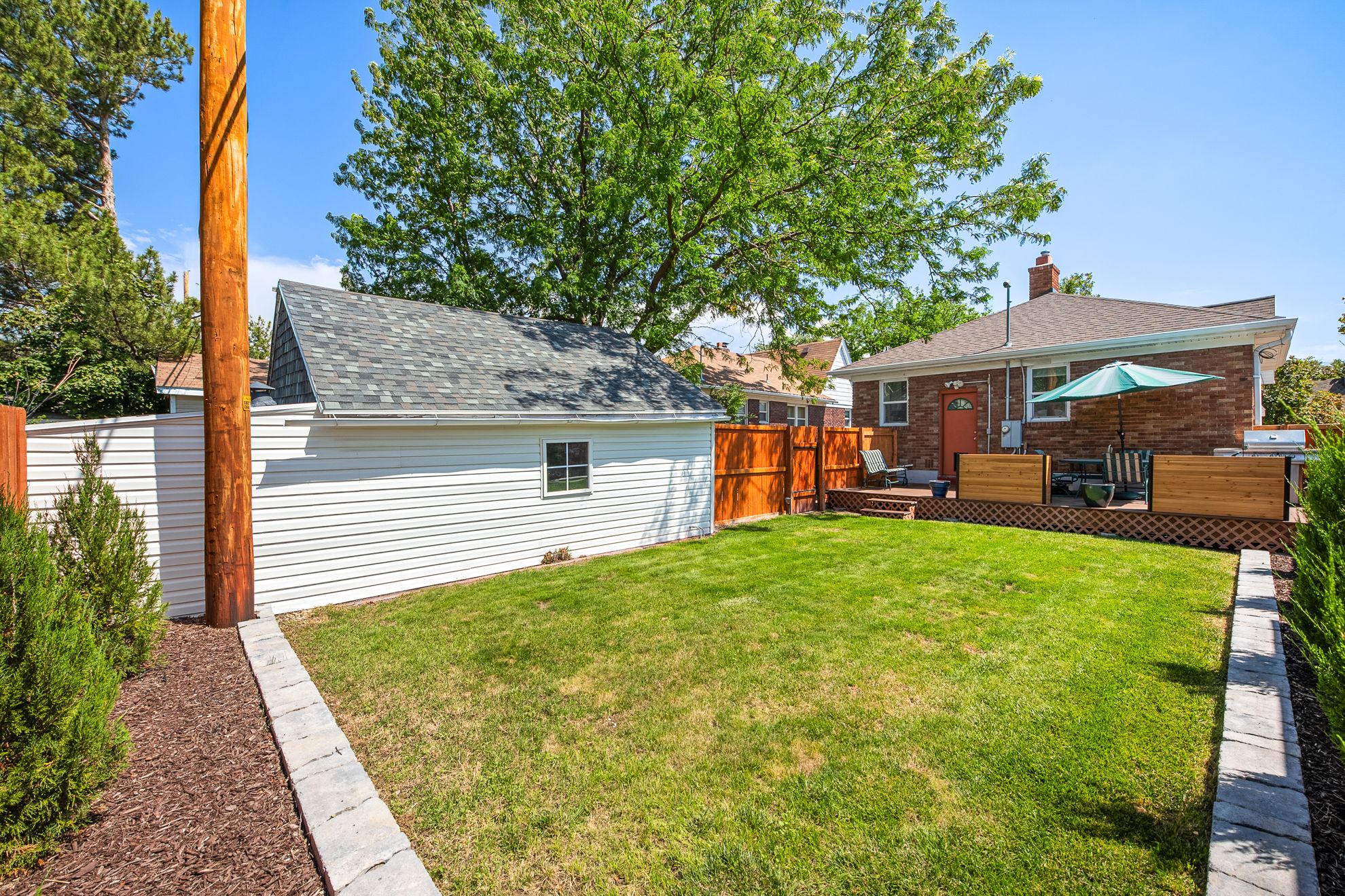 Expansive backyard featuring a manicured lawn, a detached garage with white siding and gray shingled roof, a wooden deck with slatted wood privacy panels, and a red brick exterior on the main residence