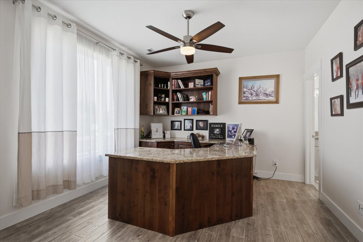 Dedicated office space featuring wood-finish flooring, a built-in wood desk with a stone countertop, built-in shelving, and a ceiling fan with integrated lighting