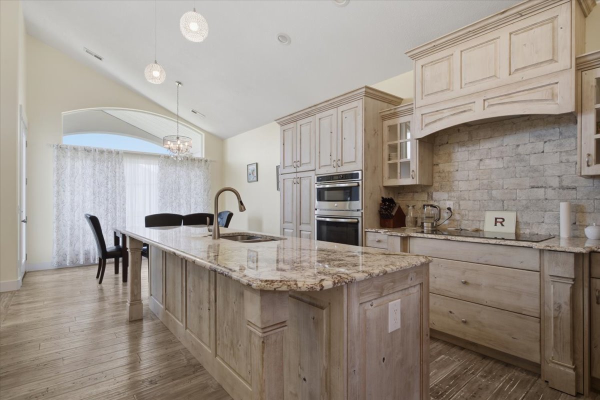 Open-concept kitchen featuring a large central island with an integrated sink, light-toned wood-finish cabinetry, a stone-finish backsplash, built-in double ovens, and wood-finish flooring