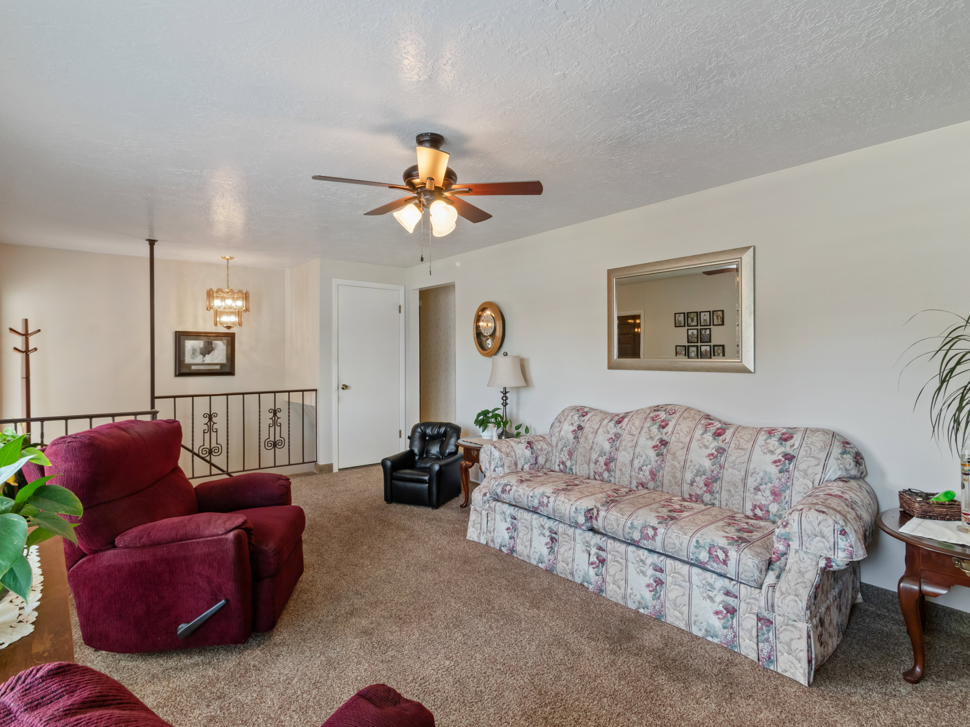 Carpeted living area featuring a ceiling fan with light fixture, white walls, and a staircase with a black metal railing