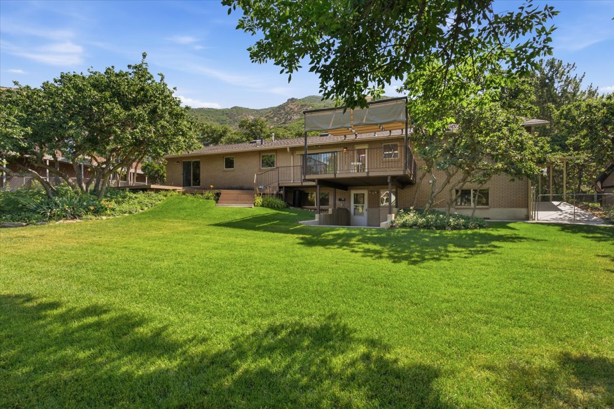 Expansive green lawn with mature trees and a two-story residence featuring a brick facade, upper-level deck with shade covering, and a lower-level patio