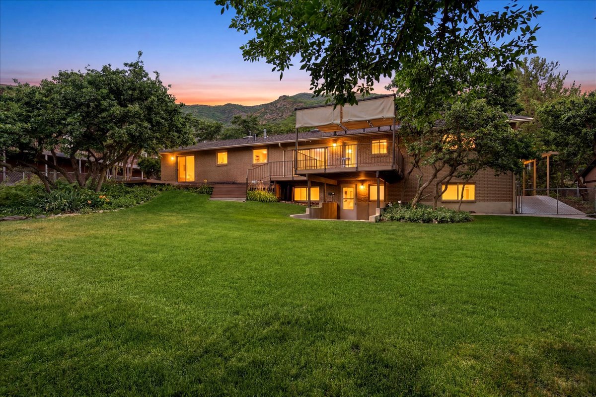 Expansive rear lawn featuring a two-story residence with a brick lower level, upper-level deck with metal railings and canopy, mature perimeter trees, and a concrete ramp structure