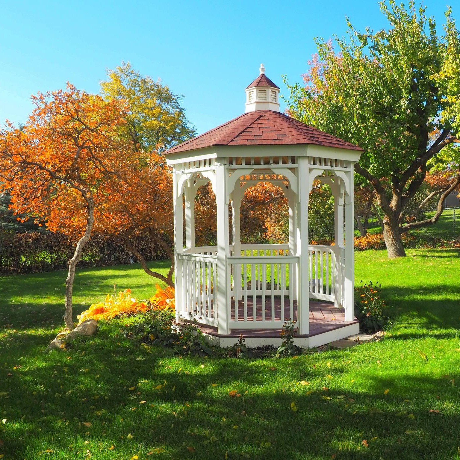 White octagonal gazebo with a red shingle roof and cupola, featuring a wood-finish floor and white baluster railing, set on a manicured lawn
