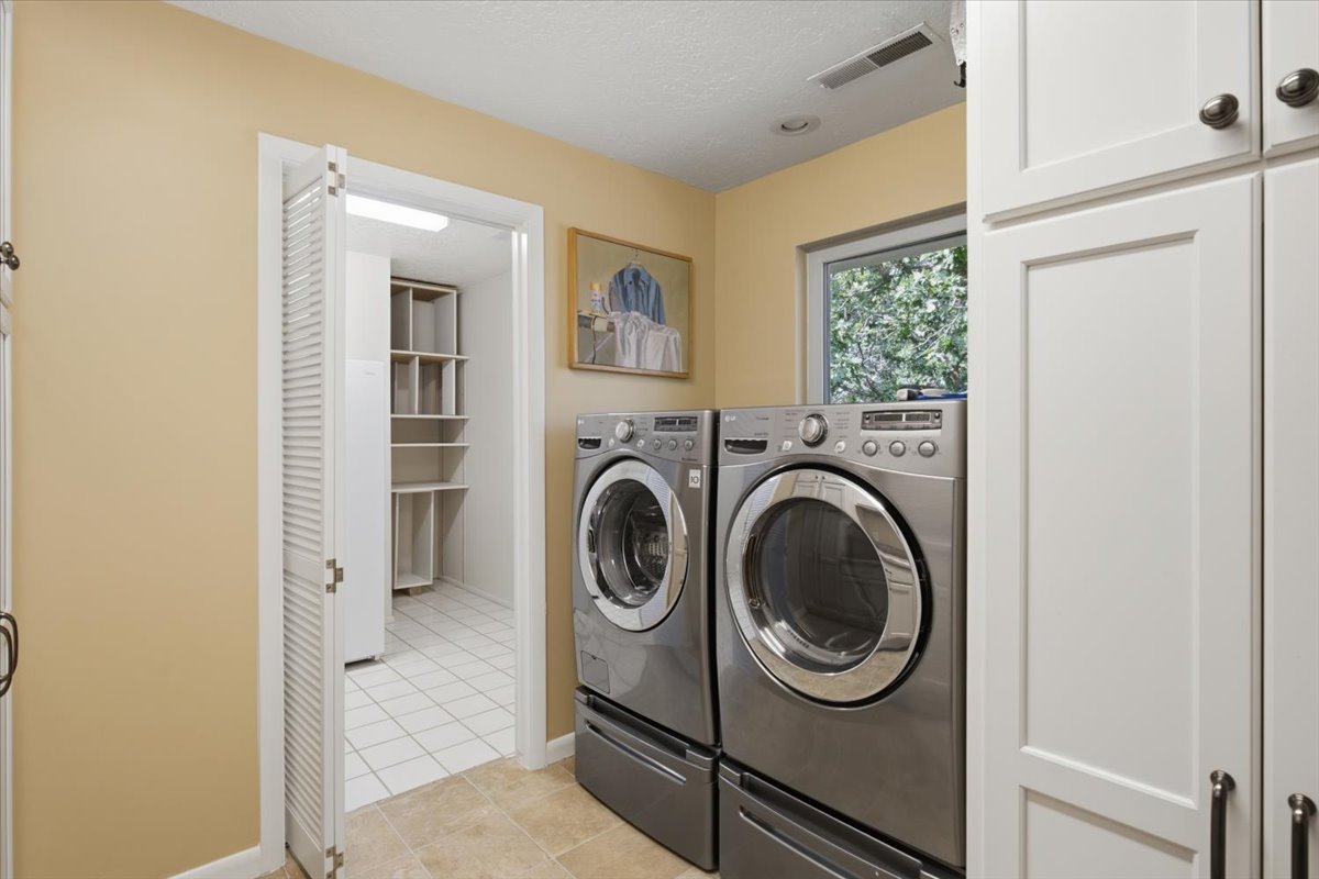 Laundry area featuring neutral wall paint, tiled flooring, white cabinetry with dark hardware, a window offering natural light, and an adjacent storage space with shelving and white tile flooring