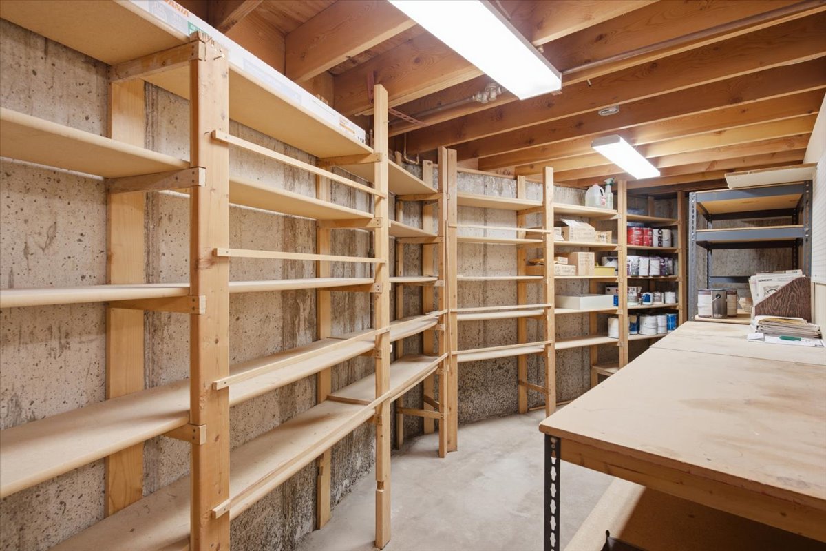 Utility space featuring extensive wood shelving, exposed concrete walls, a wood-beam ceiling, and fluorescent lighting