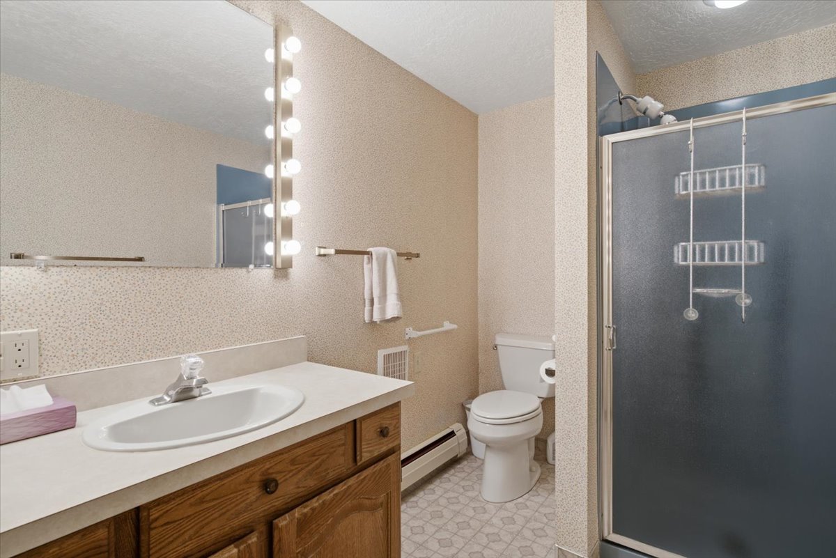 Bathroom featuring a wood-finish vanity with a white countertop and integrated sink, a large wall-mounted mirror, a vertical vanity light fixture, and a shower with a frosted glass door