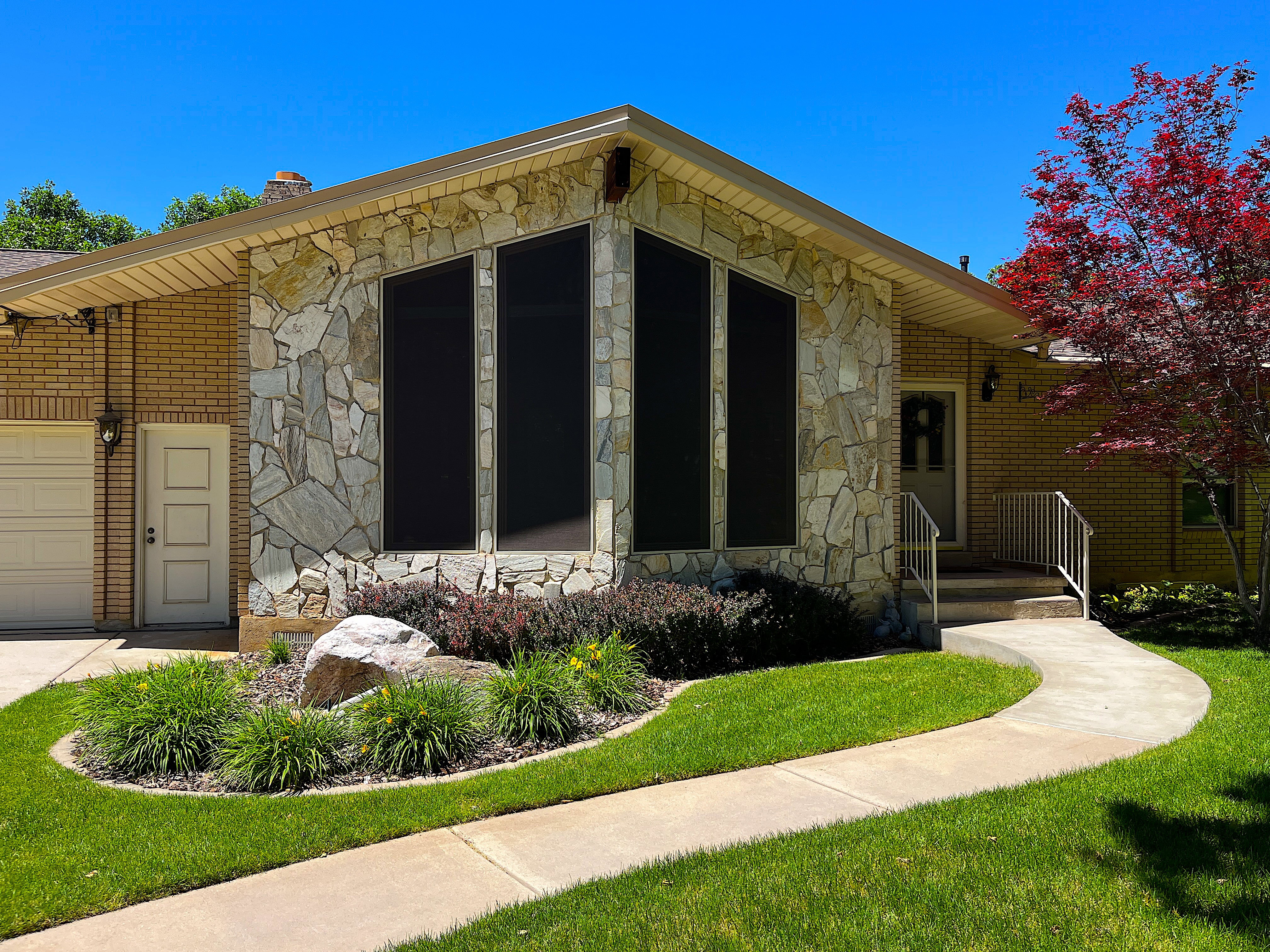 Exterior featuring a prominent stone facade, brick siding, a curved concrete pathway, a decorative rock garden, and a striking red-leafed tree