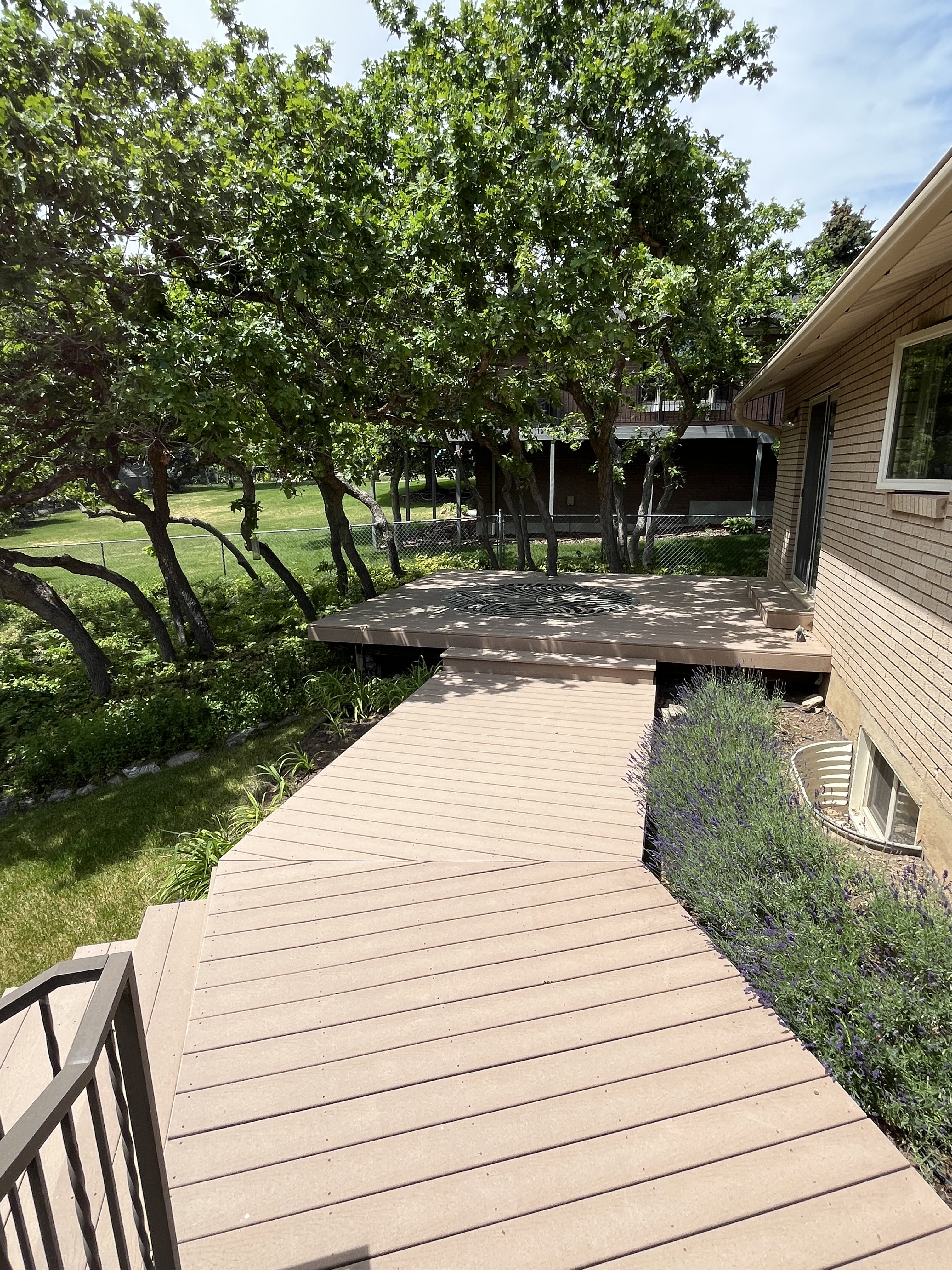 Multi-level composite decking with integrated steps, black metal railing, and mature trees providing natural shade