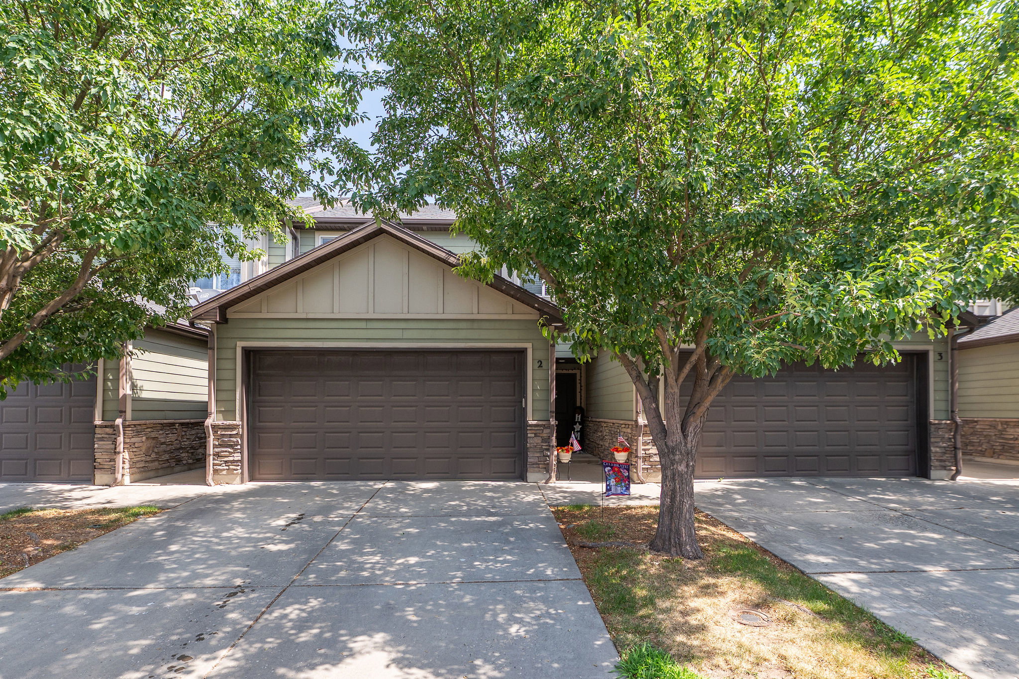 Green siding exterior with a dark brown garage door, stone accents, a concrete driveway, and mature trees