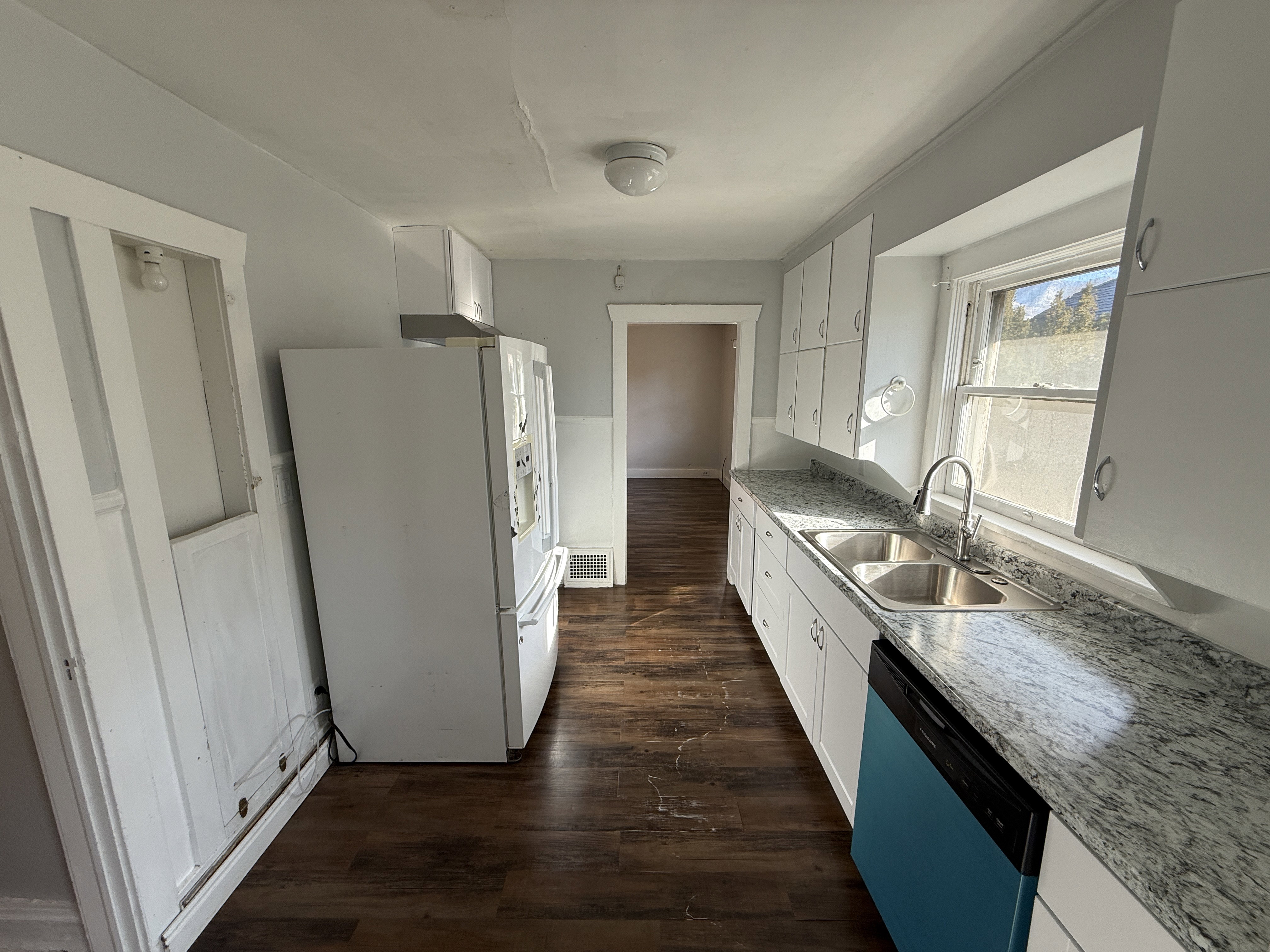 Galley kitchen featuring wood-finish flooring, white cabinetry, a double basin stainless steel sink, and light-toned countertops