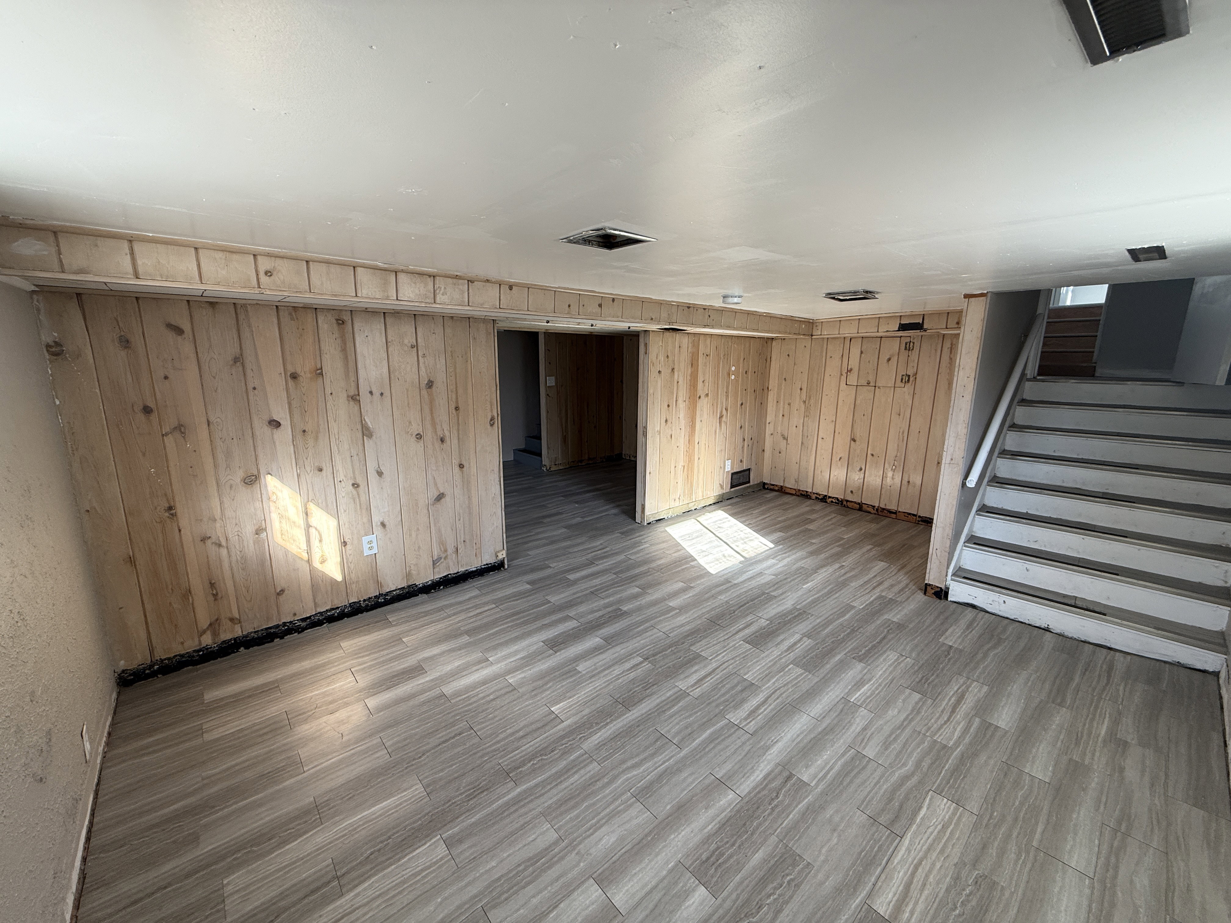 Spacious room featuring wood-finish wall paneling, light-toned tile flooring, painted ceiling, and visible stair access