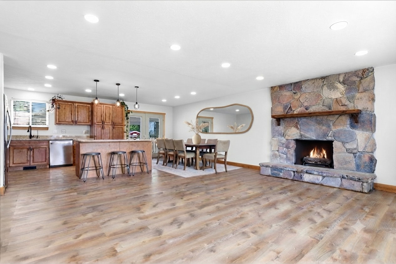 Expansive living area featuring wood-finish flooring, a stone fireplace with a substantial wood mantel, a kitchen island with pendant lighting, wood cabinetry, and stainless steel appliances