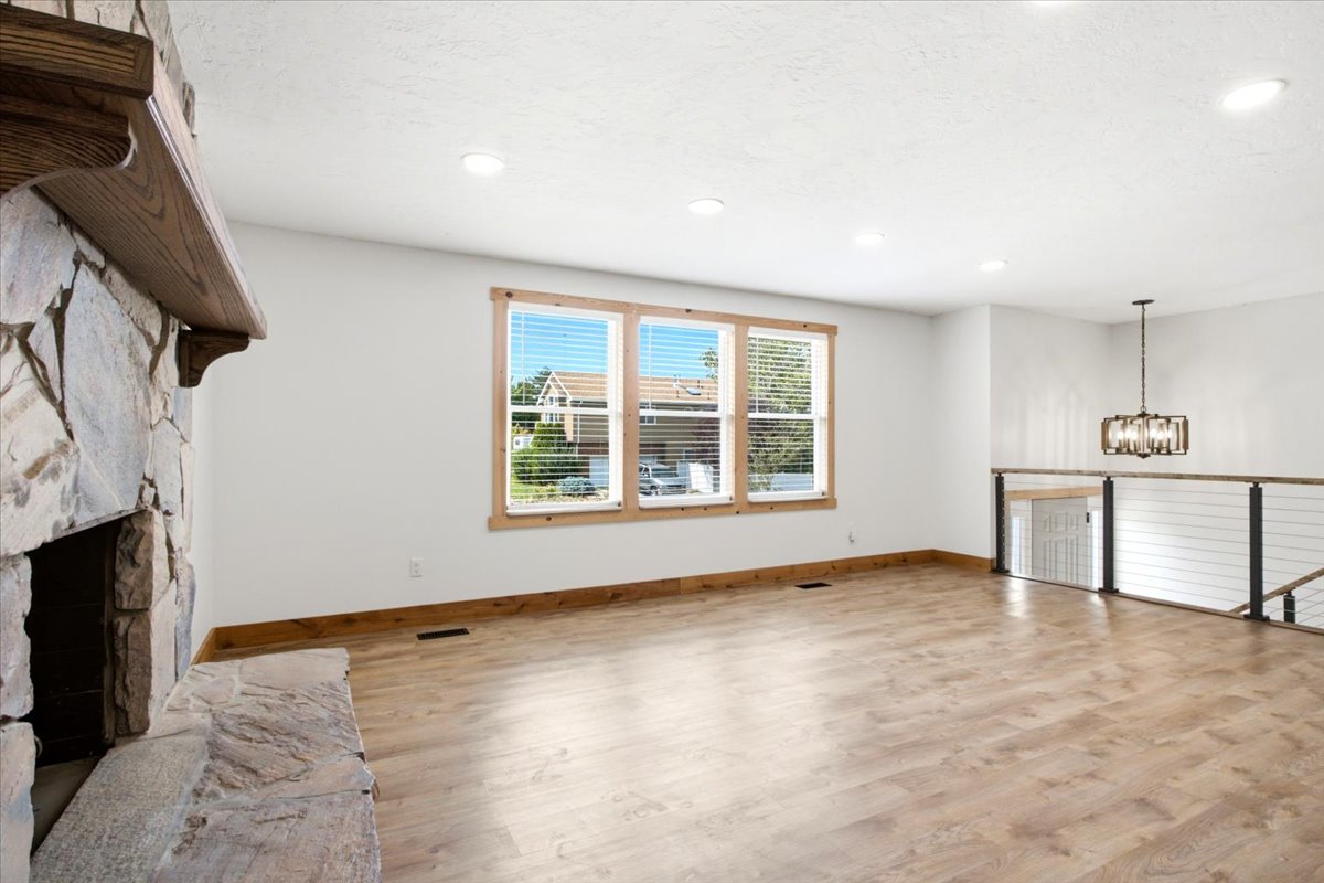 Spacious living area featuring a stone fireplace with a wood mantle, wood-finish flooring, a trio of windows with wood trim, recessed lighting, and a modern chandelier illuminating the open railing
